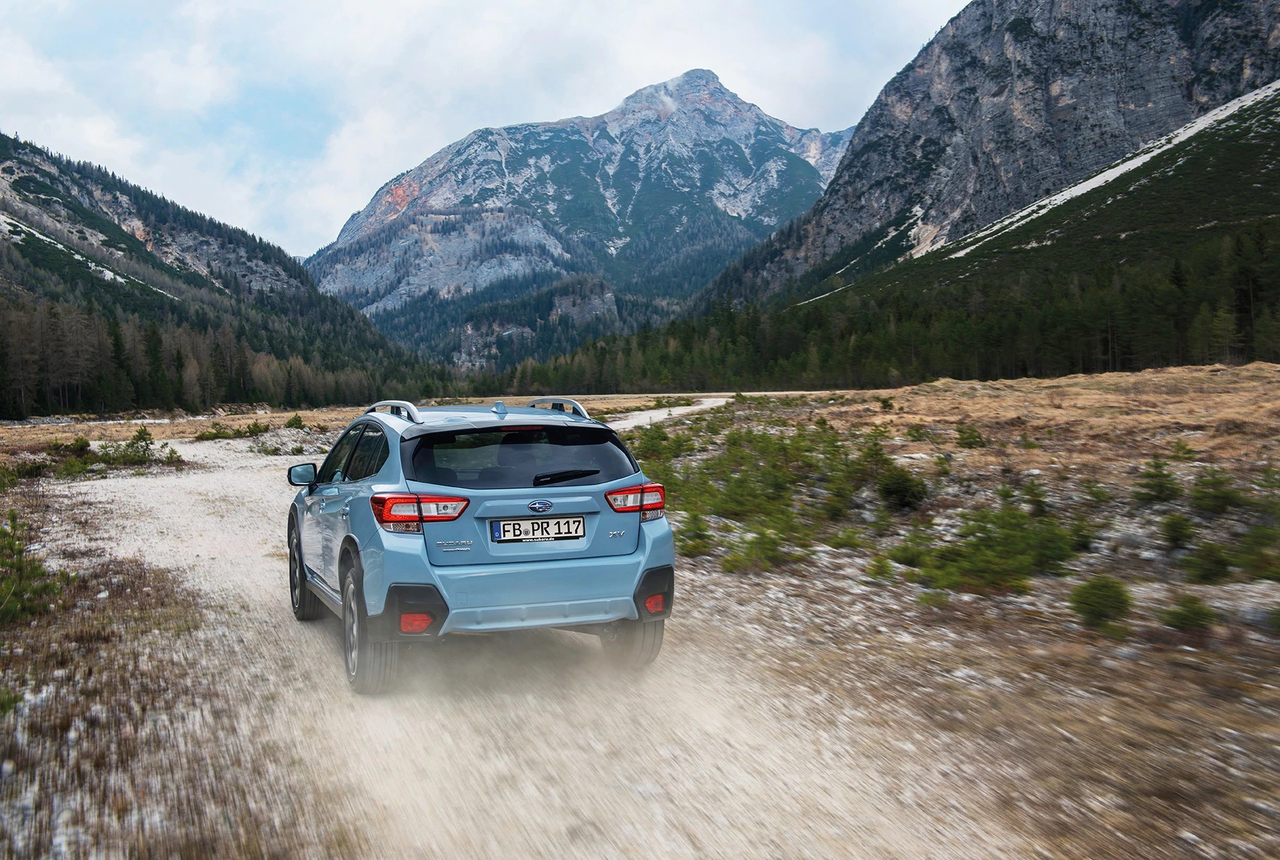 Ein blauer SUV fährt auf einem staubigen Weg durch eine Berglandschaft mit steilen Hängen und Nadelwäldern unter bewölktem Himmel.