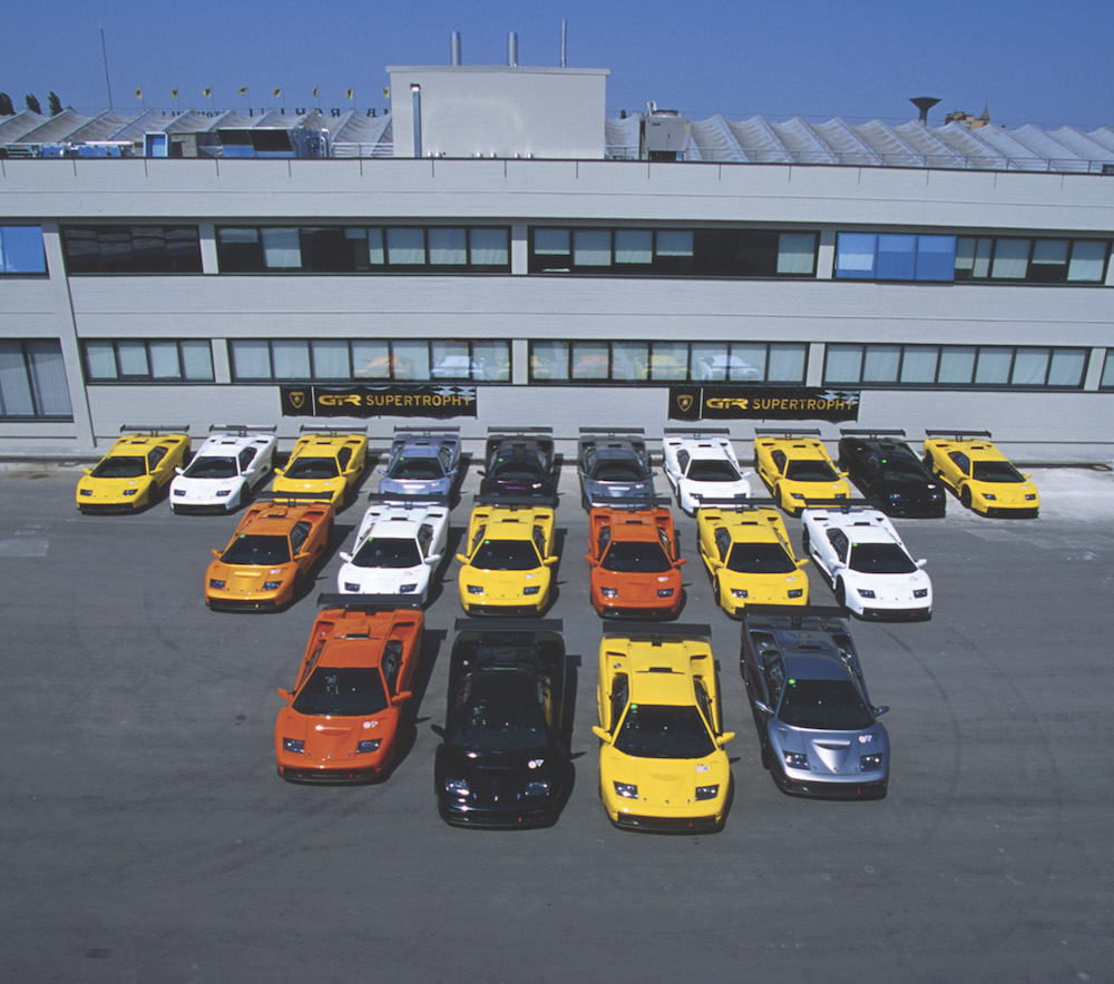 Eine Gruppe von bunten Sportwagen, nebeneinander auf einem Parkplatz vor einem grauen Gebäude in einer sonnigen Umgebung aufgestellt.