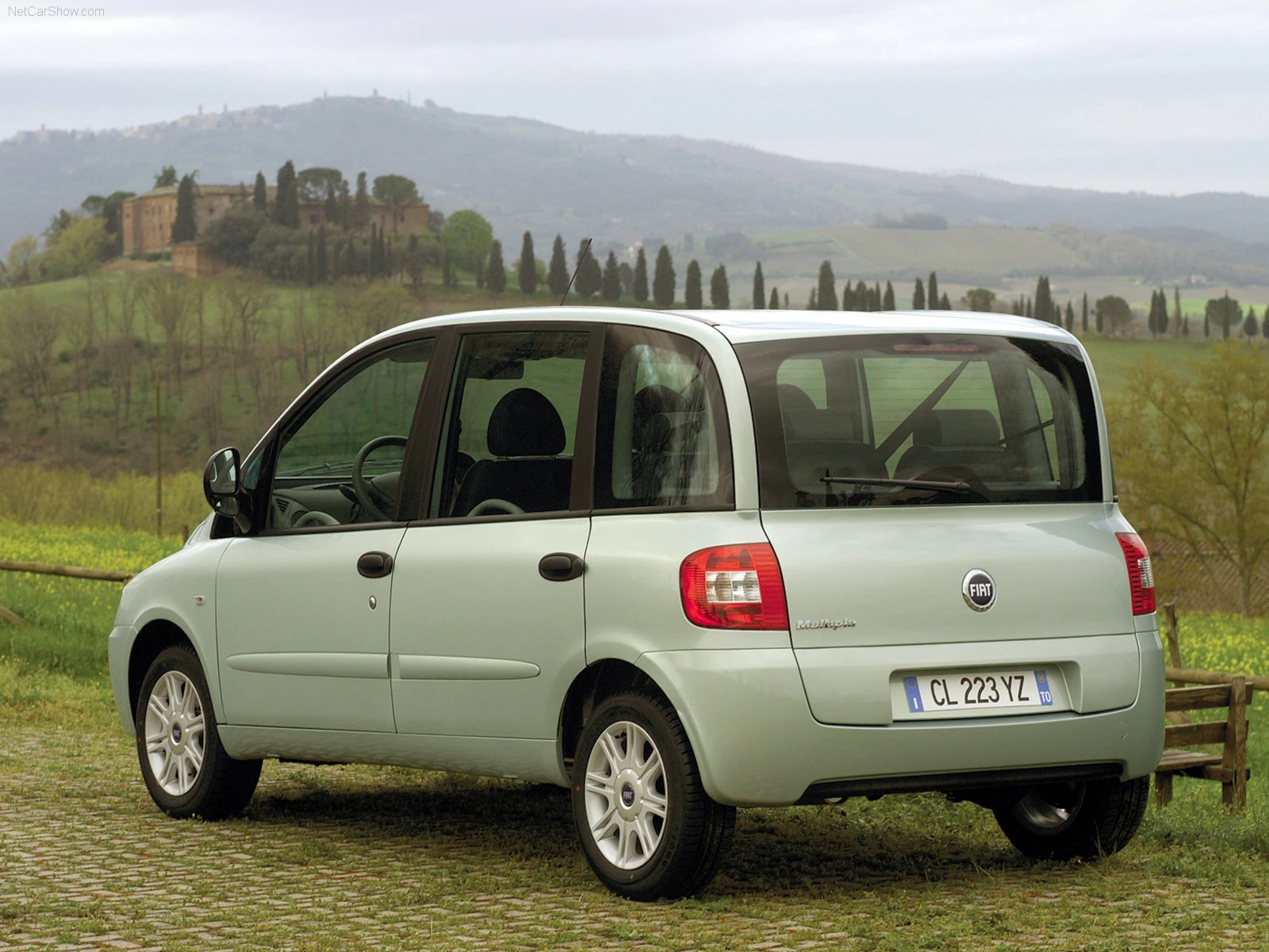 Ein hellgrüner Fiat Multipla steht auf einem Kopfsteinpflasterweg vor einer hügeligen Landschaft mit Bäumen und einem Schloss im Hintergrund.