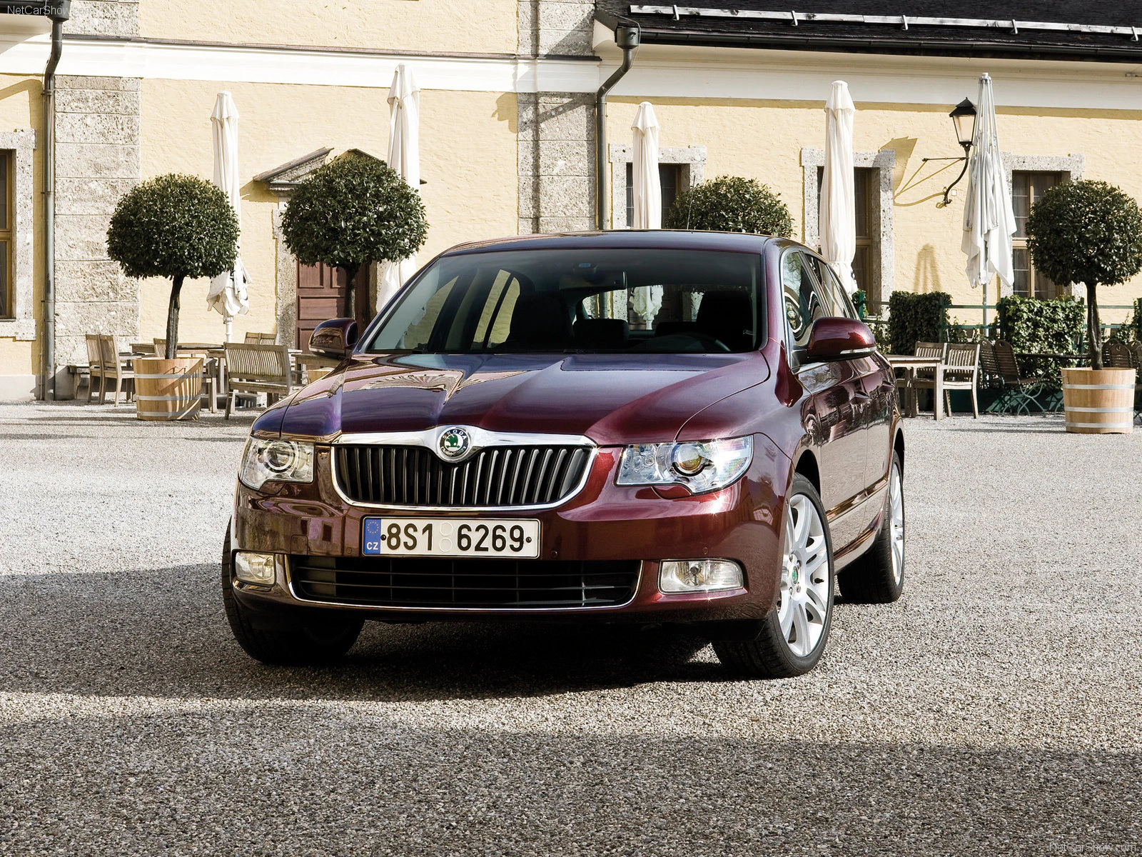 Roter Škoda Superb Limousine, geparkt auf einem Kiesplatz vor einem Gebäude mit gelben Wänden und dekorativen Bäumen im Hintergrund.