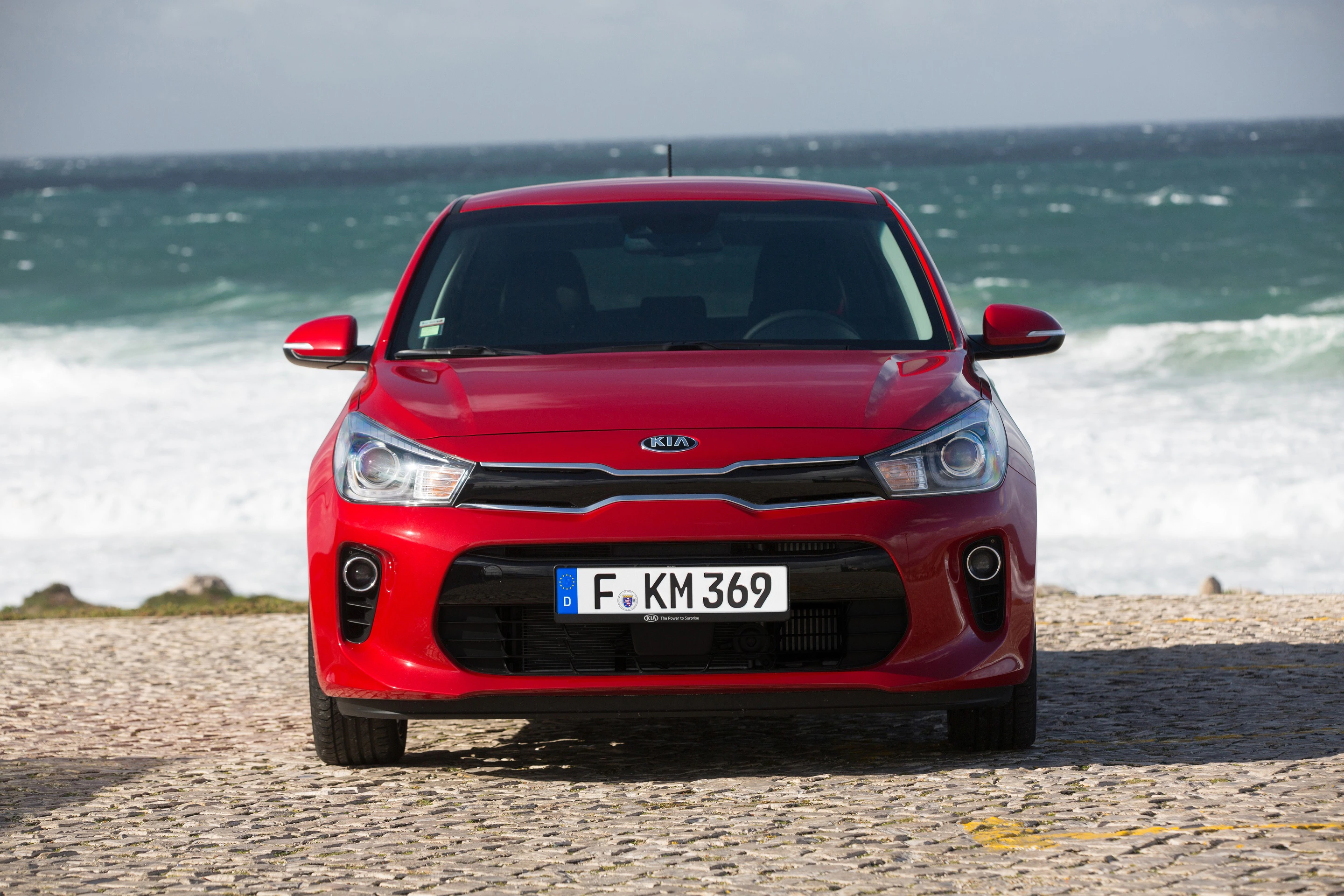 Roter Kia Rio steht auf Kopfsteinpflaster vor stürmischer See. Wellen im Hintergrund. Fahrzeug in der Mitte der Aufnahme, frontal zu sehen.
