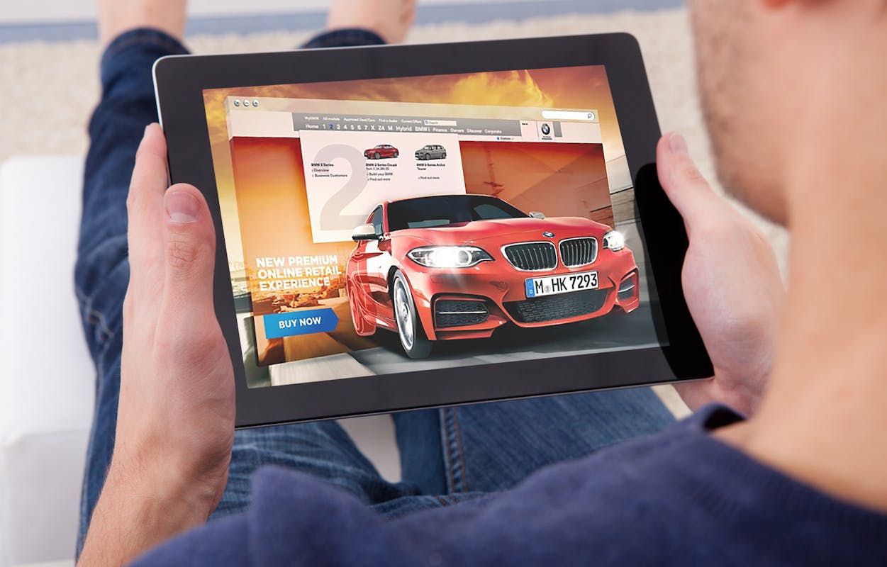 Person hält ein Tablet mit einer Webseite, die ein rotes Auto und einen "Buy Now"-Button zeigt, auf dem Schoß.