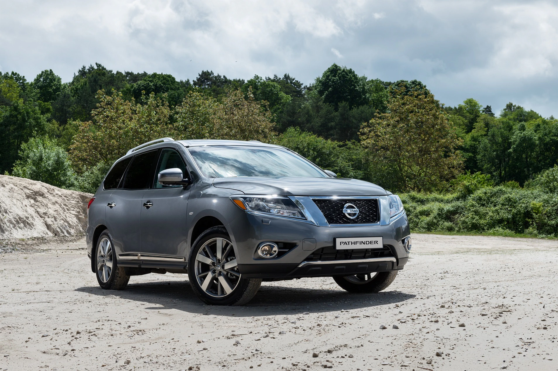 Grauer SUV der Marke Nissan steht auf einer sandigen Fläche vor einem Hintergrund aus Bäumen und bewölktem Himmel.