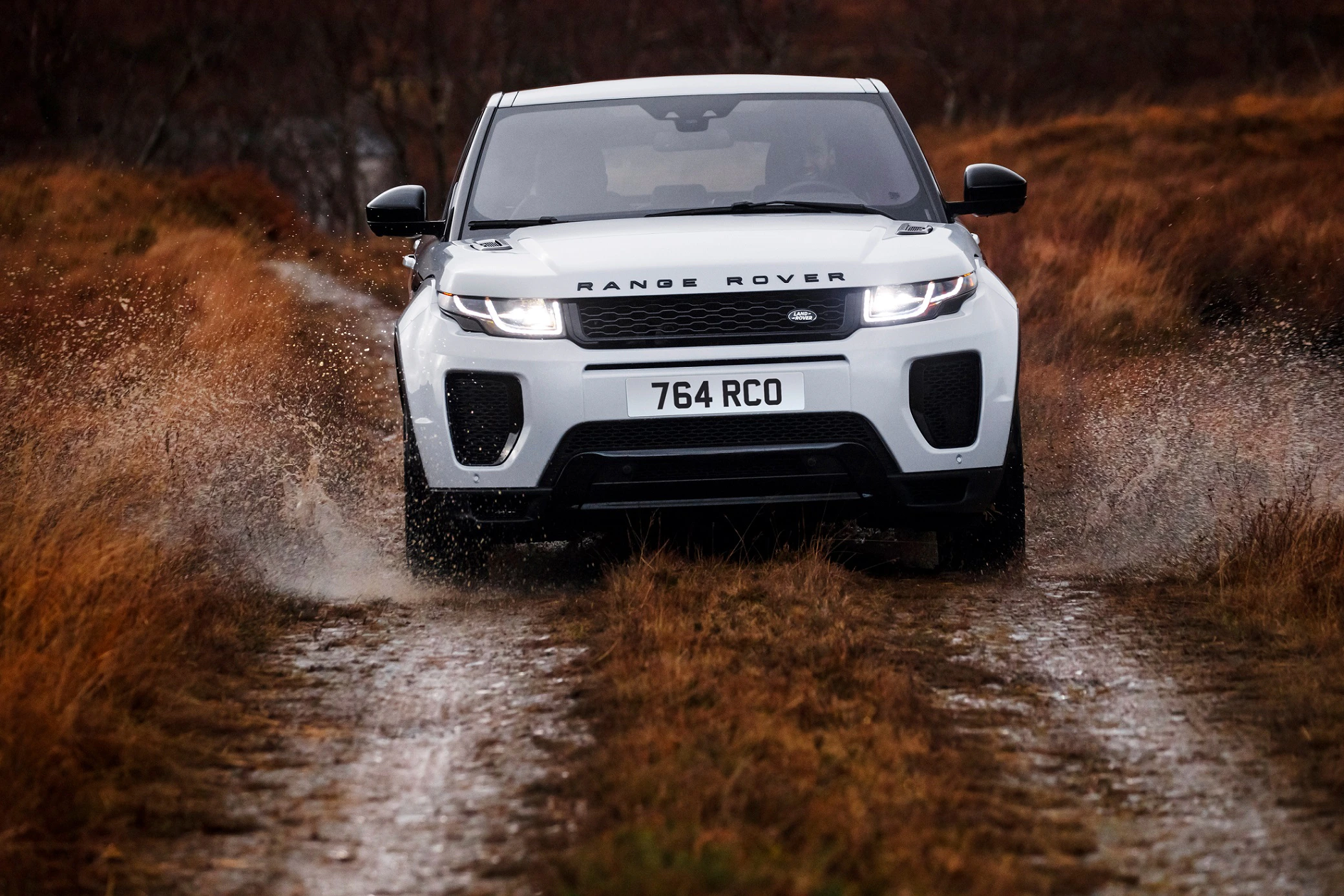 Ein weißer Range Rover fährt schnell durch eine schlammige, wasserbedeckte Pfütze auf einem ländlichen Weg.