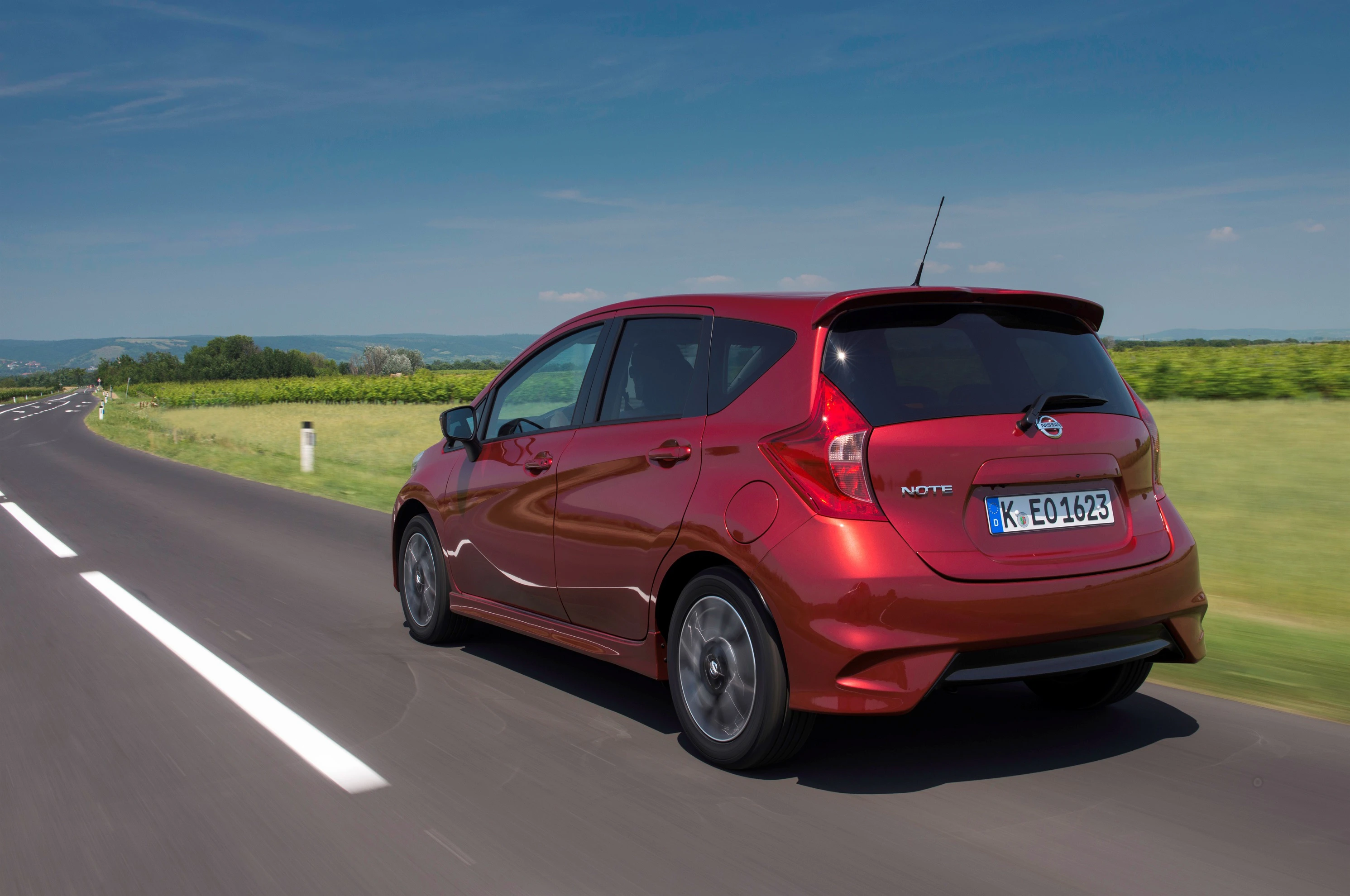 Rotes Auto auf einer Landstraße, umgeben von grünen Feldern und blauem Himmel. Das Fahrzeug fährt in Richtung Horizont.