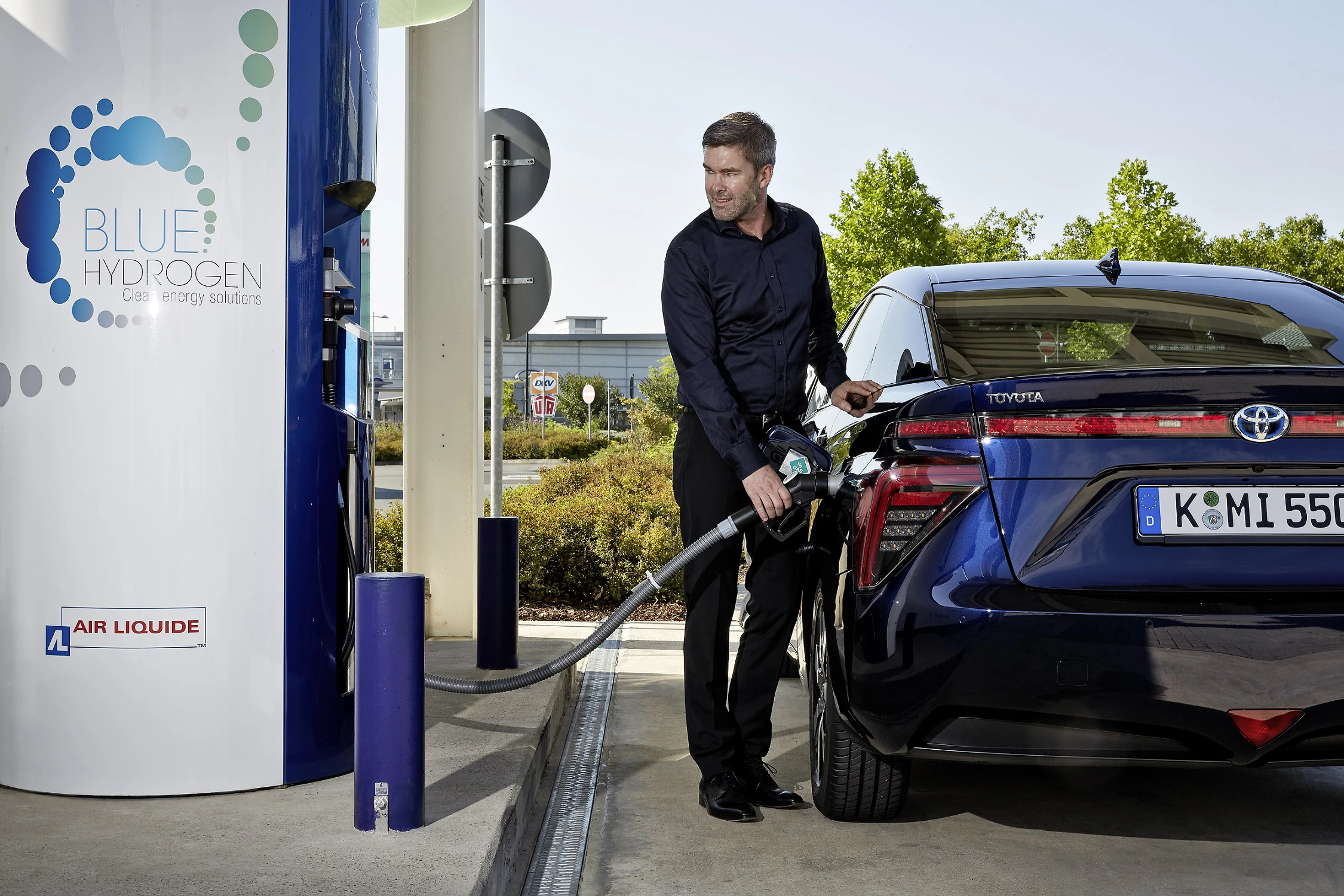 Ein Mann betankt ein blaues Auto mit Wasserstoff an einer Tankstelle. Im Hintergrund Bäume und ein Gebäude.