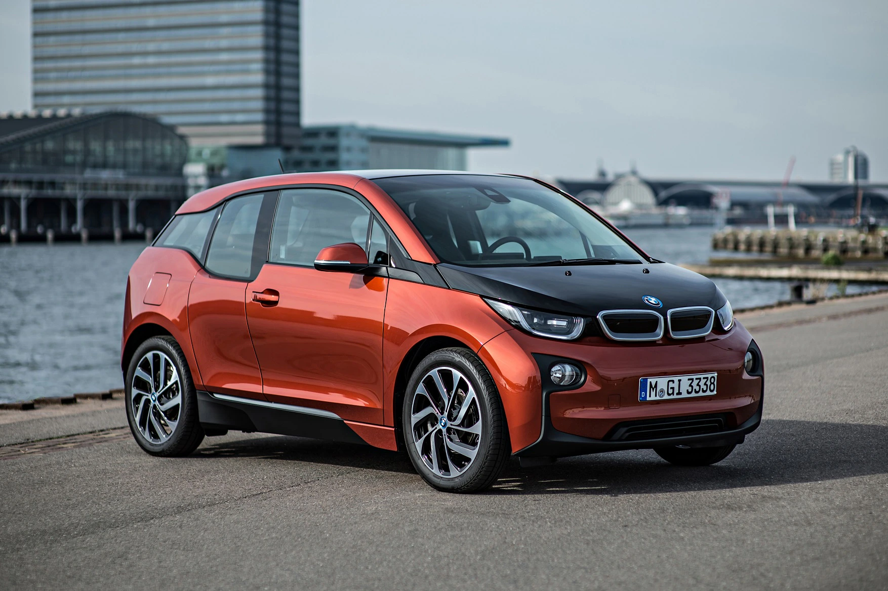 Roter BMW i3 steht auf einer Straße am Wasser. Hintergrund: modernes Gebäude, Himmel bewölkt. Elektrischer Kleinwagen mit futuristischem Design.