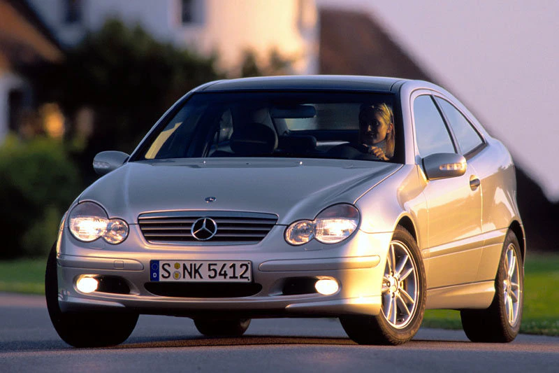 Silberner Mercedes-Benz C-Klasse Coupé fährt auf einer Straße bei Tageslicht. Frau am Steuer, Hintergrund verschwommen.