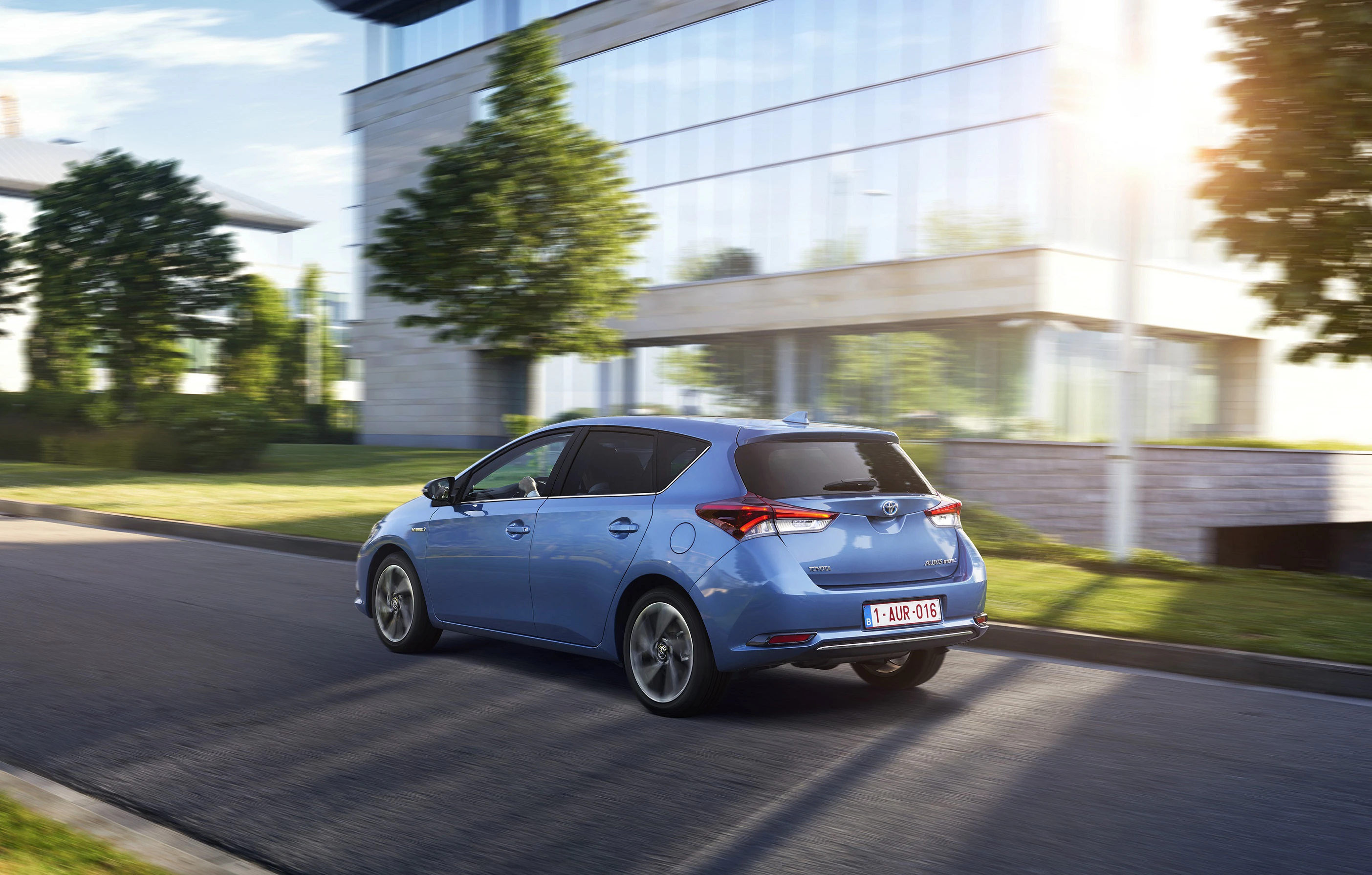 Blaues Auto fährt auf einer Straße neben einem modernen Gebäude mit Glasfassade, umgeben von Bäumen und Sonnenlicht.