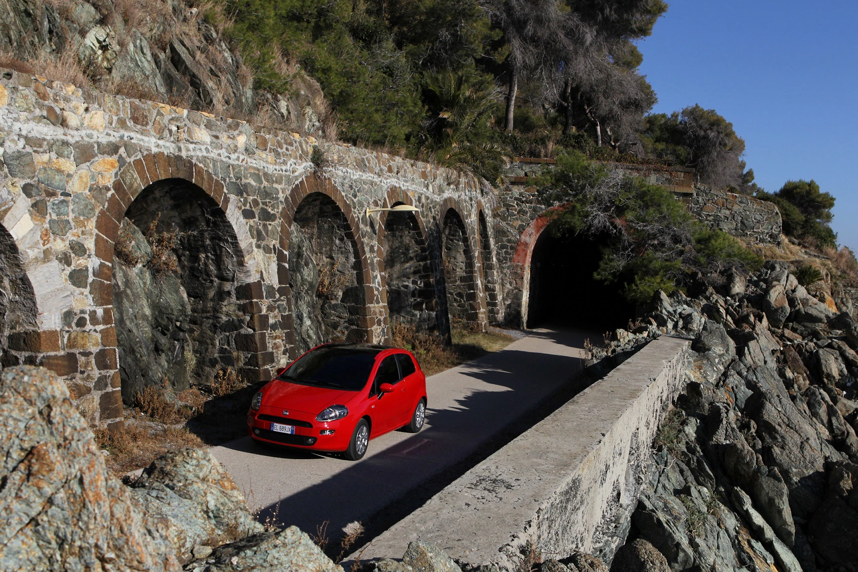 Rotes Auto auf schmaler Straße neben historischen Steinbögen und Felsen, führt in einen Tunnel. Sonniges Wetter, klare, blaue Himmelsansicht.