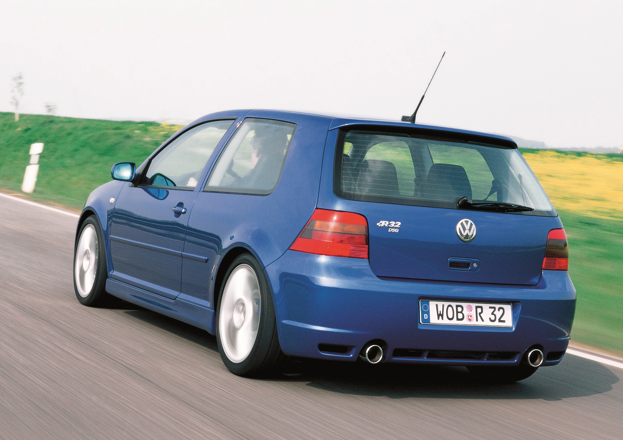 Blauer VW Golf R32 fährt auf einer Landstraße. Rückansicht mit charakteristischen Doppelauspuffrohren und sportlichem Design.