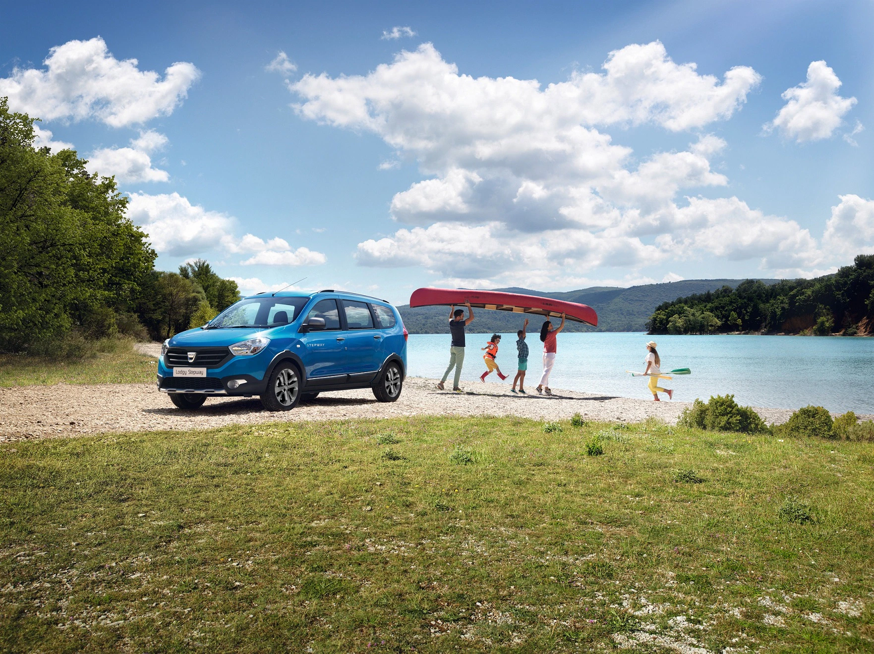 Blaues Auto am Seeufer. Eine Familie trägt ein rotes Kanu zum Wasser. Sonniger Tag, Wolken am Himmel.