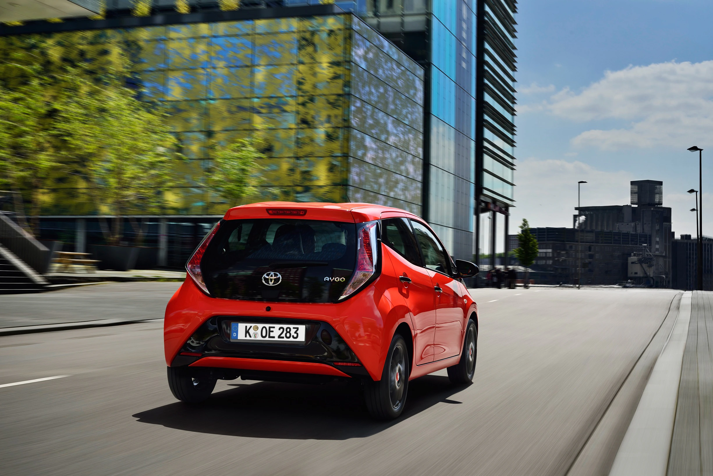 Roter Toyota Aygo fährt auf leerer urbaner Straße, moderne Gebäude im Hintergrund, sonniges Wetter.