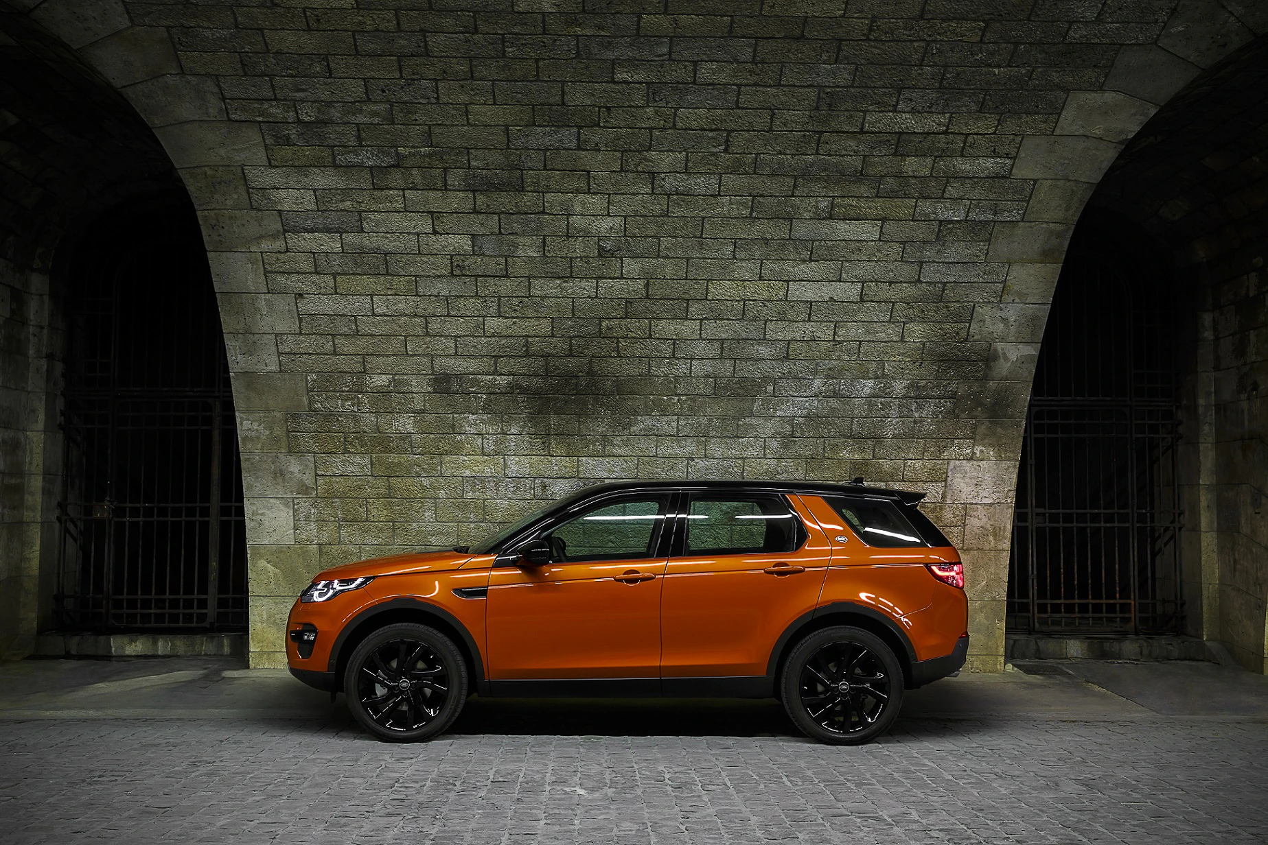 Ein orangefarbener SUV parkt vor einer gewölbten Steinwand, mit eleganten schwarzen Felgen und dunklen Fenstern.