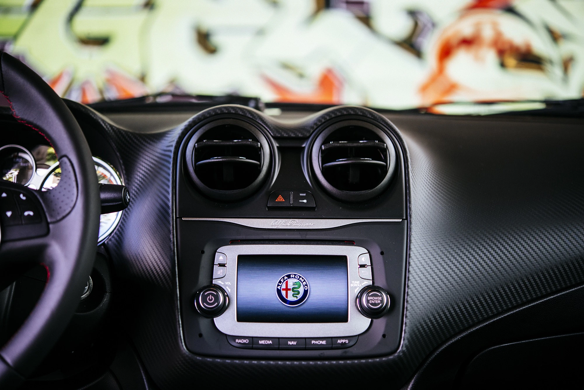 Nahaufnahme eines Autoinnenraums mit einem Bildschirm, der das Alfa Romeo-Logo zeigt, umgeben von Lüftungsöffnungen und Steuerknöpfen.