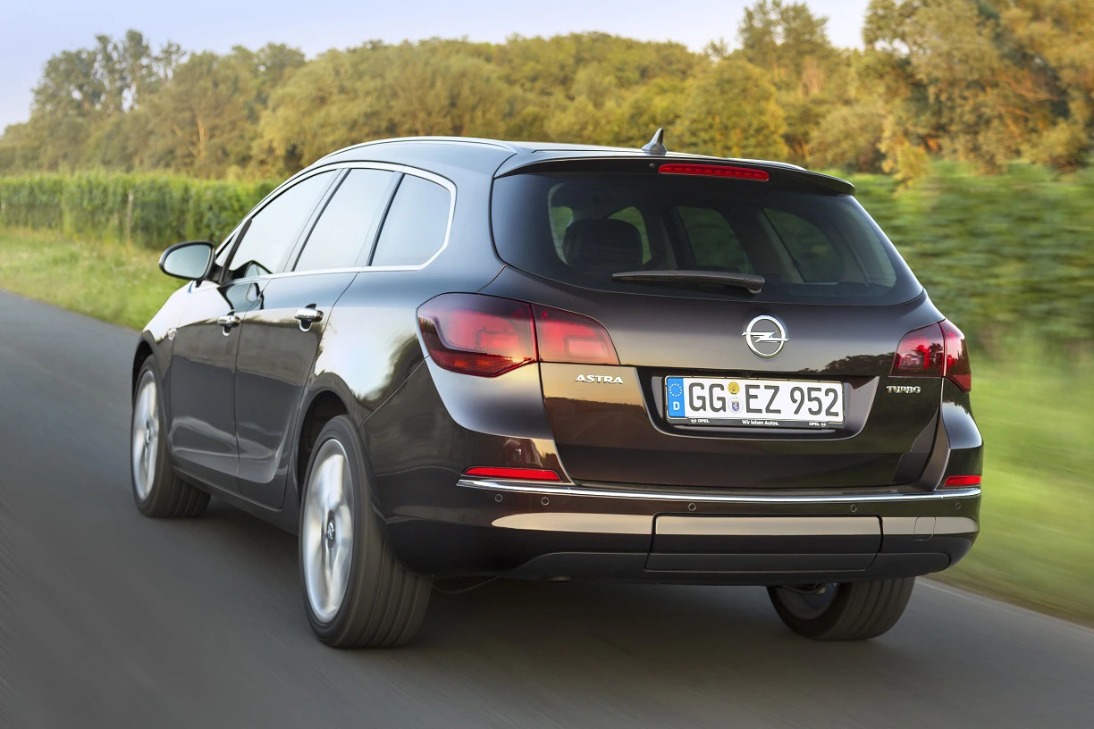 Ein dunkelbrauner Opel Astra Sports Tourer fährt auf einer Landstraße, umgeben von Bäumen und Feldern im Hintergrund.