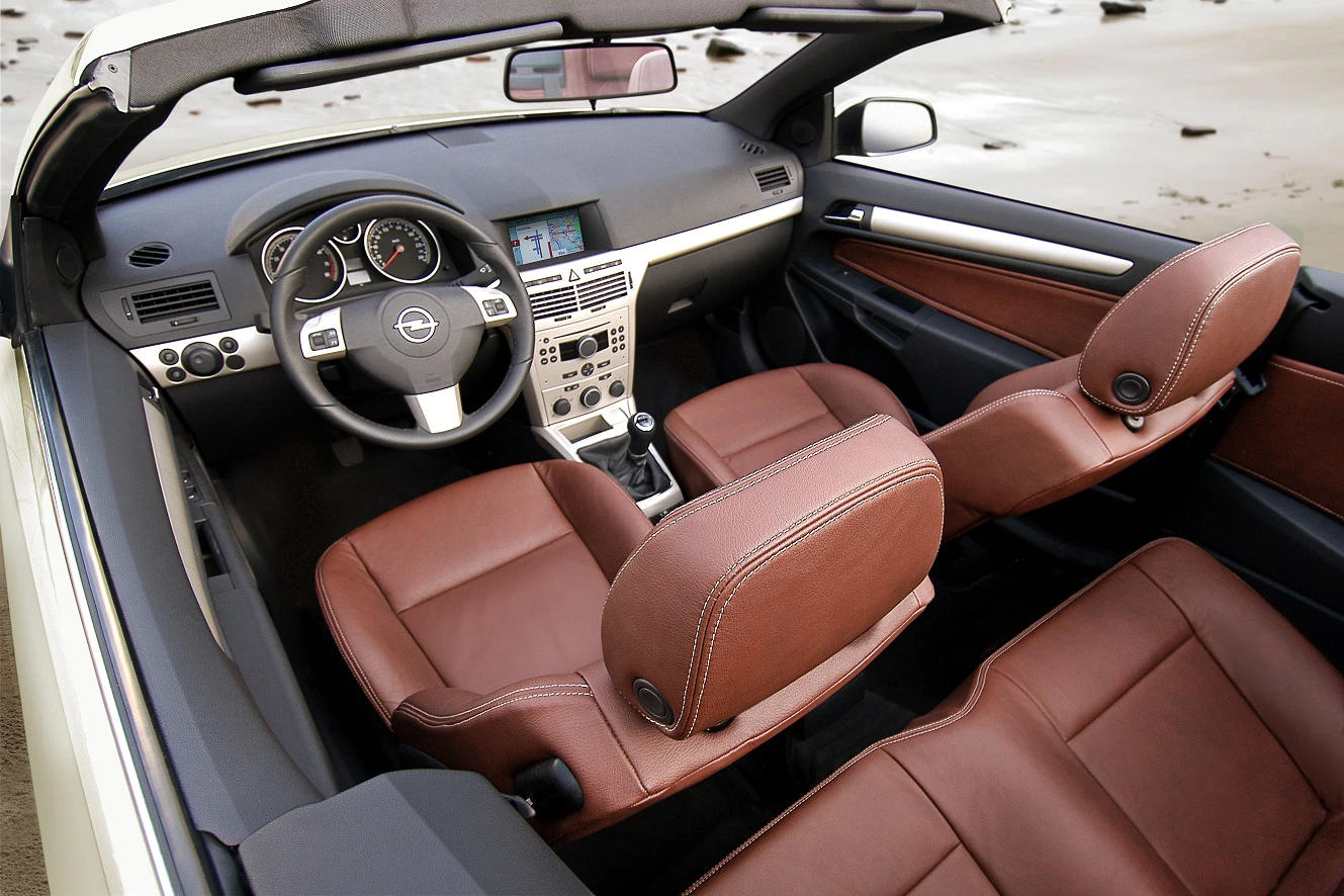 Innenraum eines Cabriolets mit braunen Ledersitzen, sportlichem Lenkrad und modernem Armaturenbrett. Der Strand ist im Hintergrund sichtbar.