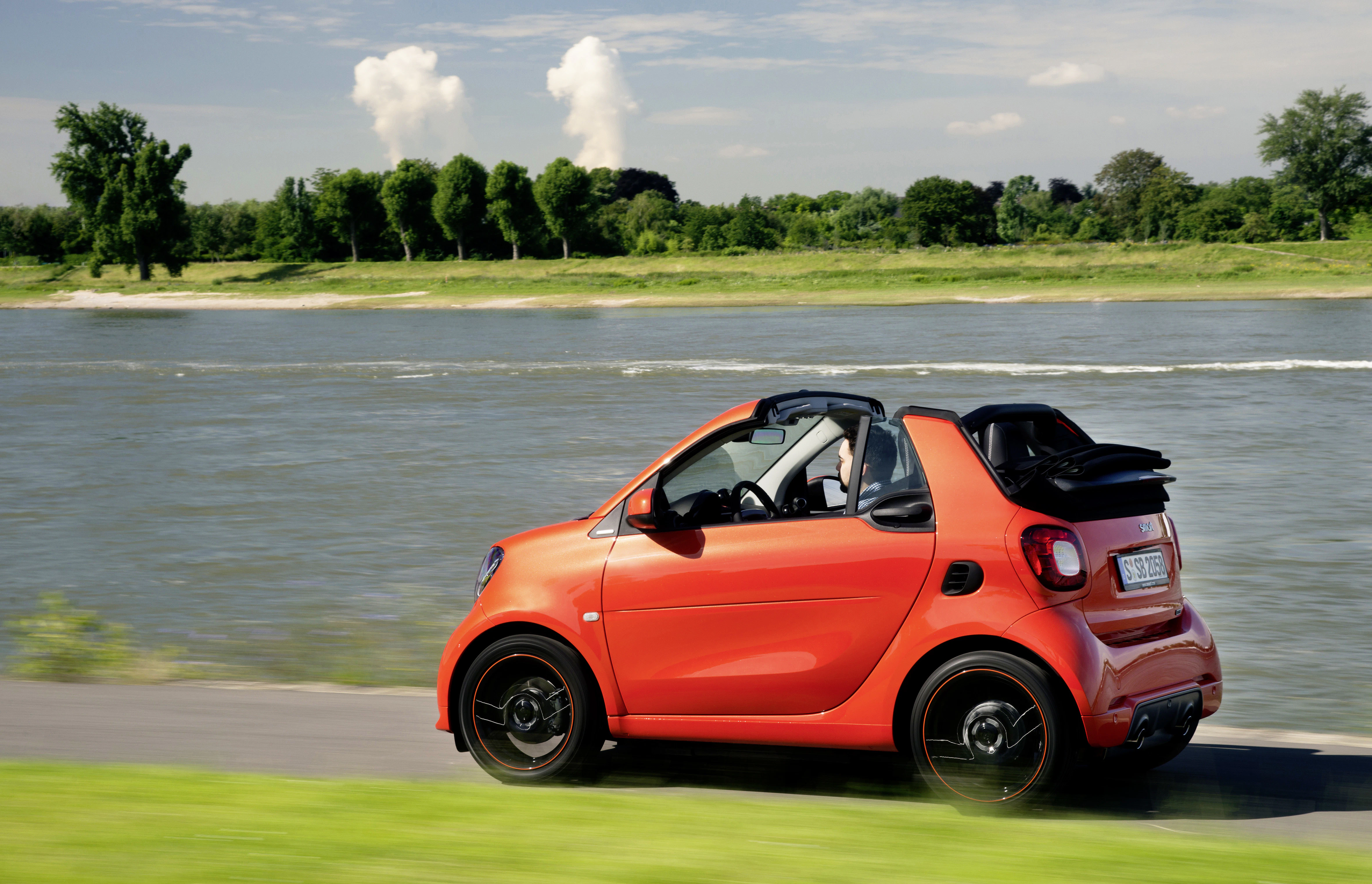 Rotes Smart-Cabrio fährt an einem Fluss entlang, bei schönem Wetter mit grünem Ufer und blauem Himmel im Hintergrund.