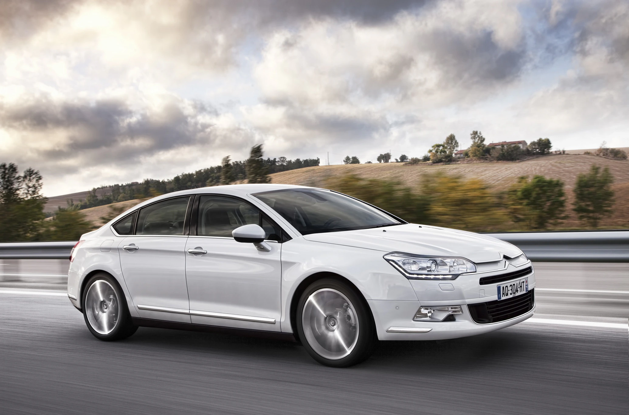 Weißer Citroën C5 Limousine fährt auf einer Landstraße bei bewölktem Himmel, mit verschwommenen Bäumen im Hintergrund.