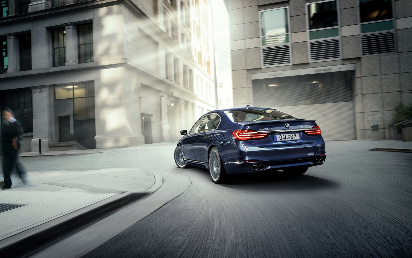 Blauer BMW fährt durch eine urbane Straßenszene bei Tageslicht, umgeben von hohen Gebäuden. Lichtstrahlen fallen auf die Straße.