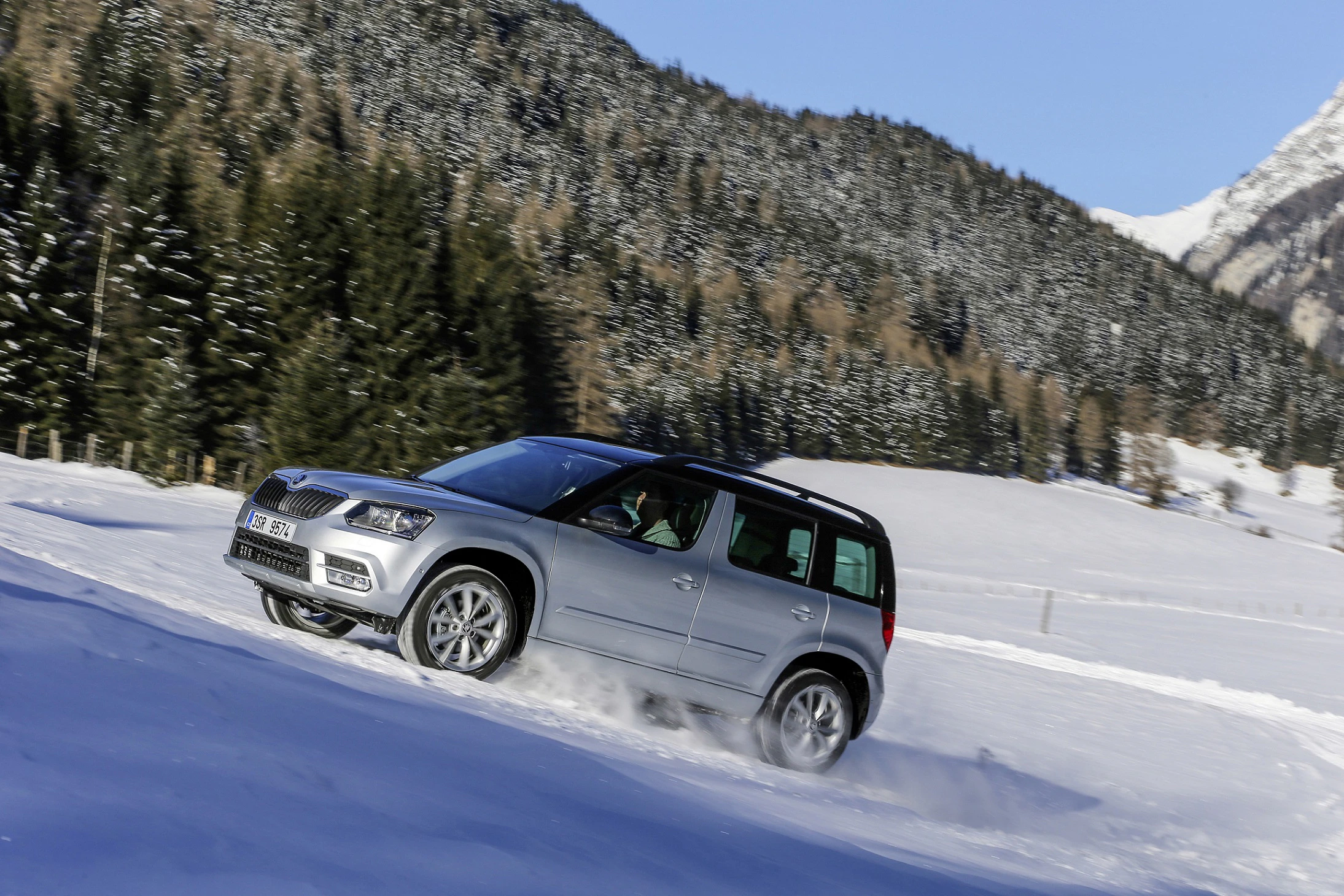 Silberner SUV fährt auf schneebedecktem Hang, umgeben von Nadelbäumen und Bergen im Hintergrund. Klarer Himmel.