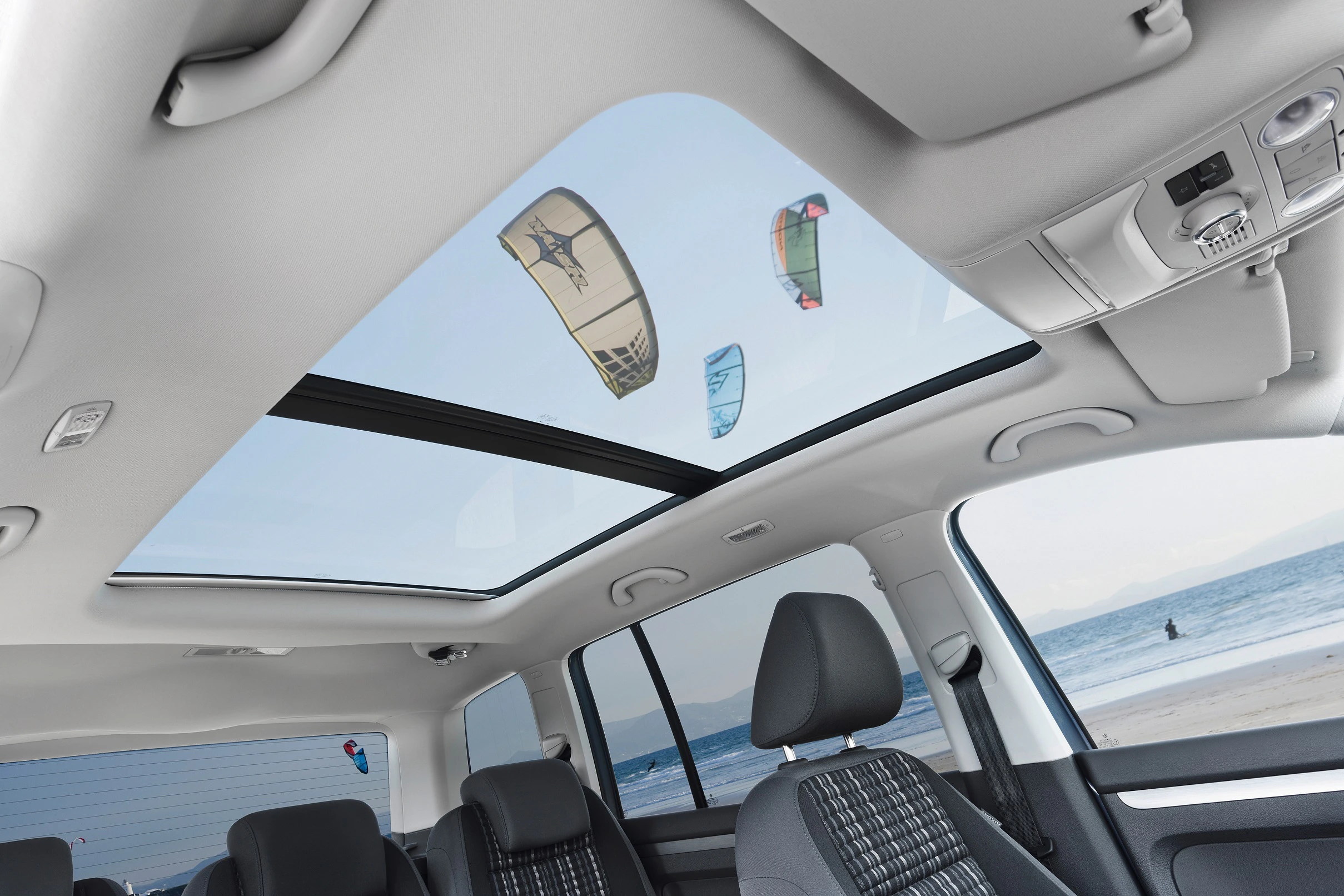Innenraum eines Autos mit Panoramadach, durch das man Kites am blauen Himmel und einen Strand sieht.