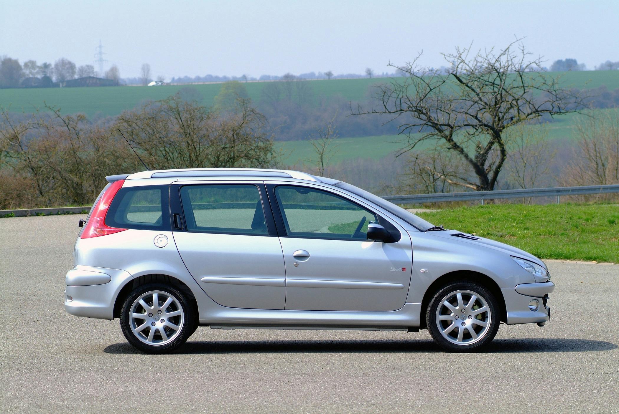 Silberner Kombi parkt auf einer Straße, umgeben von kahlen Bäumen und grüner Landschaft unter blauem Himmel.