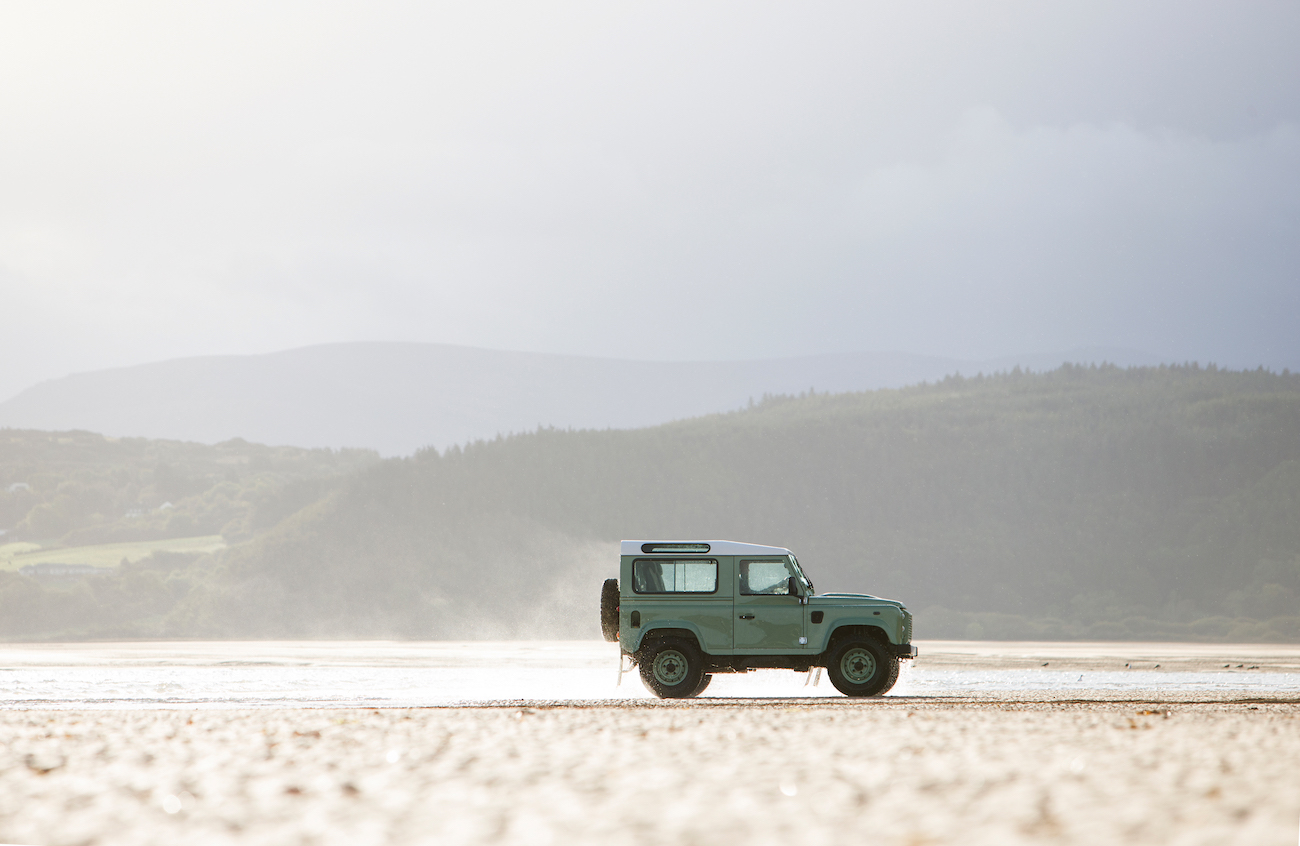 Ein grüner Geländewagen fährt über einen weiten, sandigen Strand vor einer hügeligen Landschaft unter bewölktem Himmel.