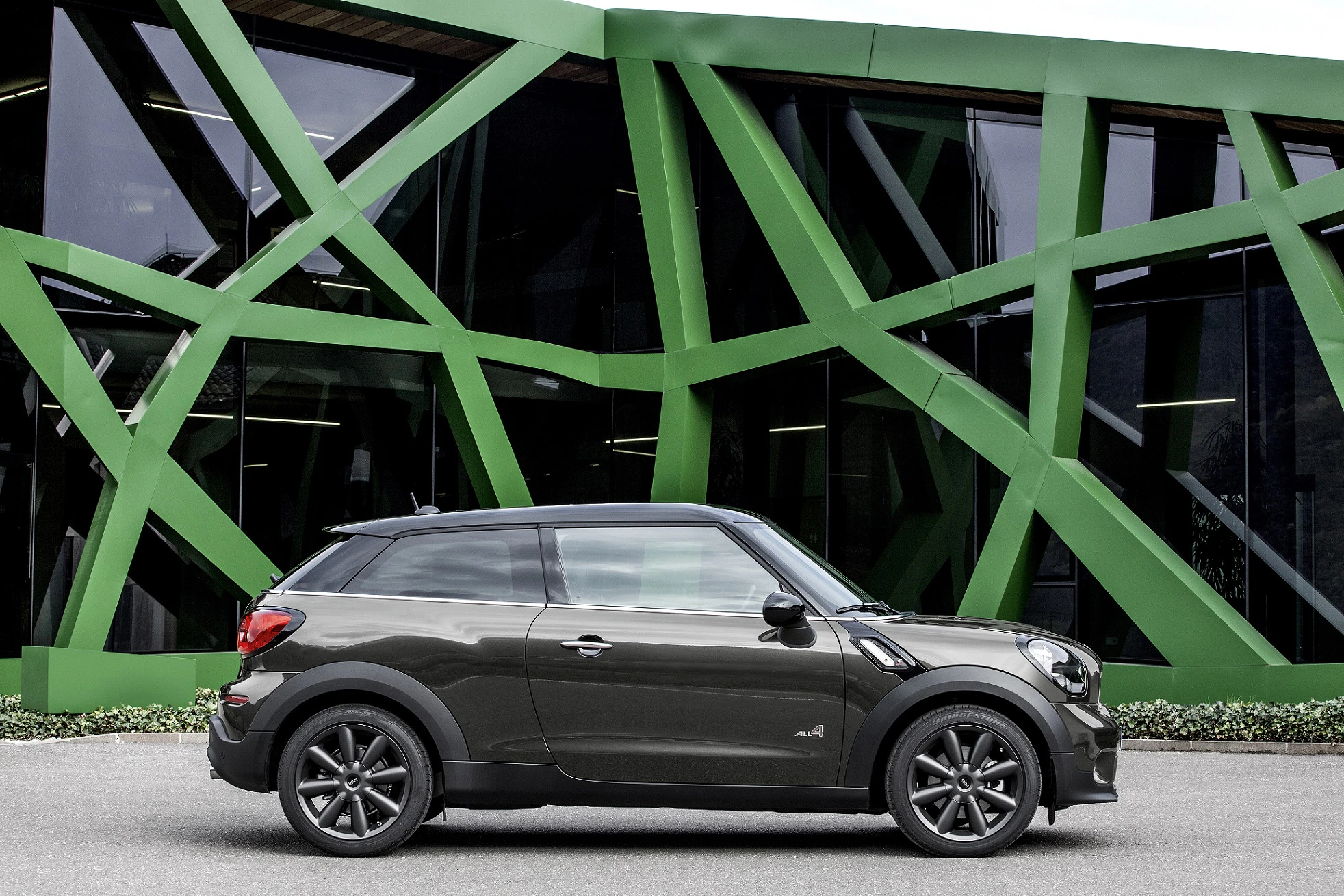 Schwarzer Mini Cooper vor modernem Gebäude mit grünen, diagonalen Stützstrukturen und großen Glasfenstern.