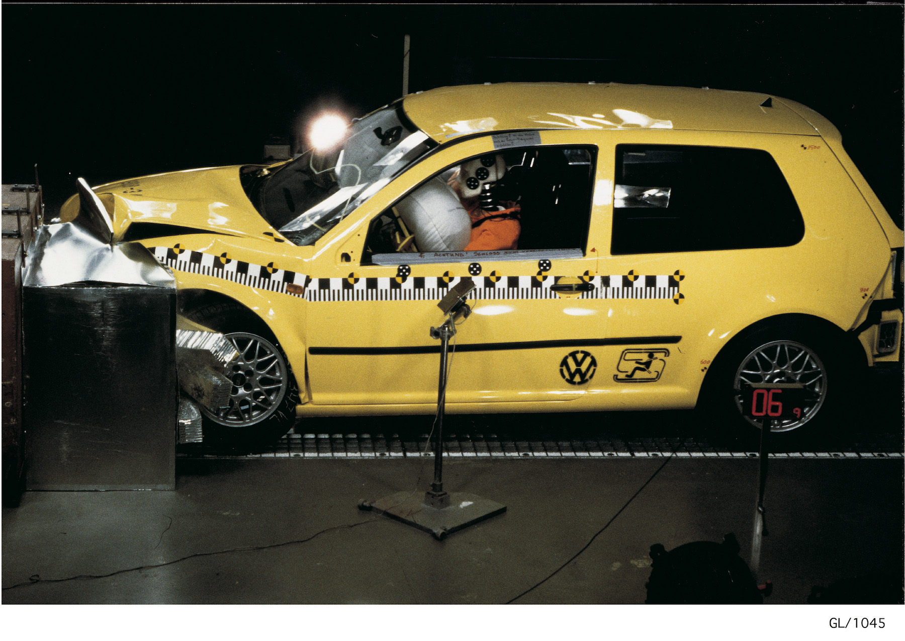 Gelber VW Golf bei einem Crash-Test, Stoßdämpfer verformt, Front kollidiert mit Barriere, Sicherheitsdummys sitzen innen.