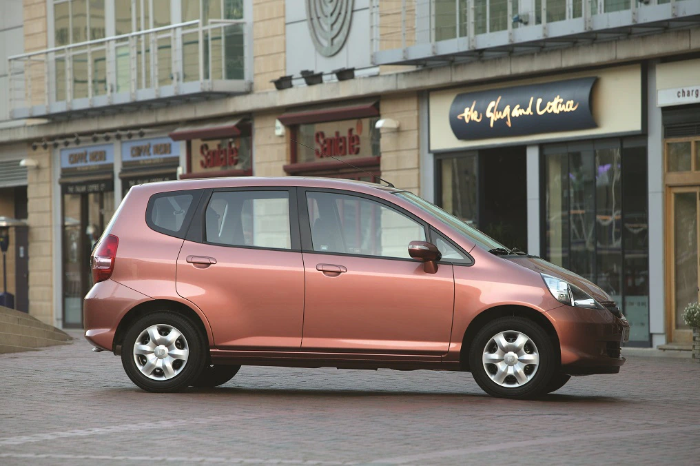 Kupferfarbener Kleinwagen steht vor modernen Geschäften mit großen Fenstern und Beschriftungen auf einem gepflasterten Platz.