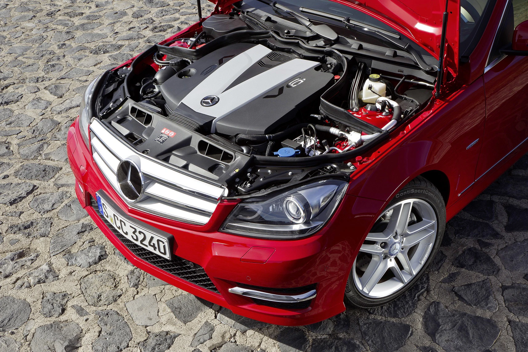 Roter Mercedes mit geöffneter Motorhaube, zeigt den Motor auf einem Kopfsteinpflaster. Frontscheinwerfer und Logo sind sichtbar.