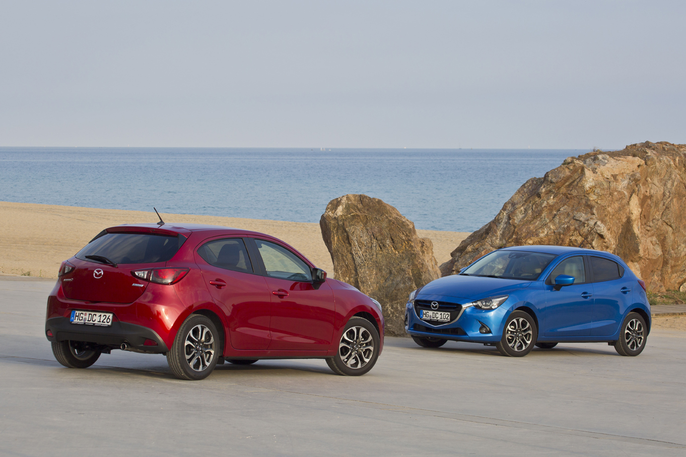 Zwei Mazda-Autos, ein rotes und ein blaues, parken vor einem felsigen Strand mit blauem Meer im Hintergrund.