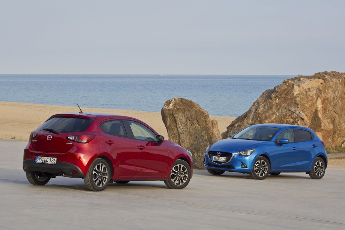 Zwei Mazda-Autos, ein rotes und ein blaues, parken vor einem felsigen Strand mit blauem Meer im Hintergrund.