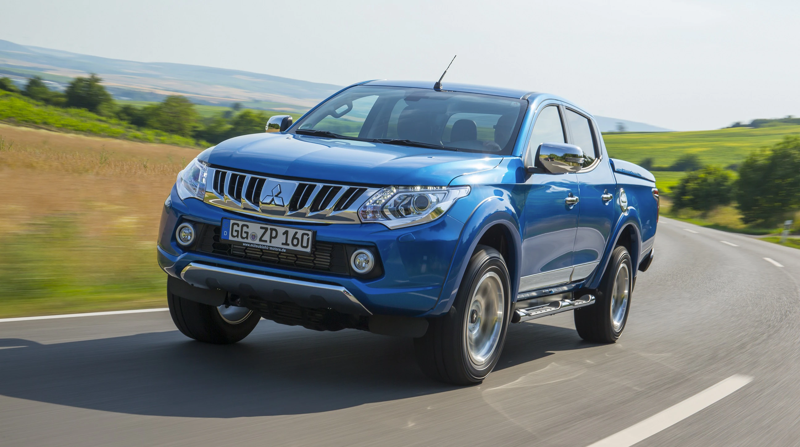 Blauer Mitsubishi-Pickup fährt auf einer Landstraße durch eine grüne Landschaft bei klarem Himmel.