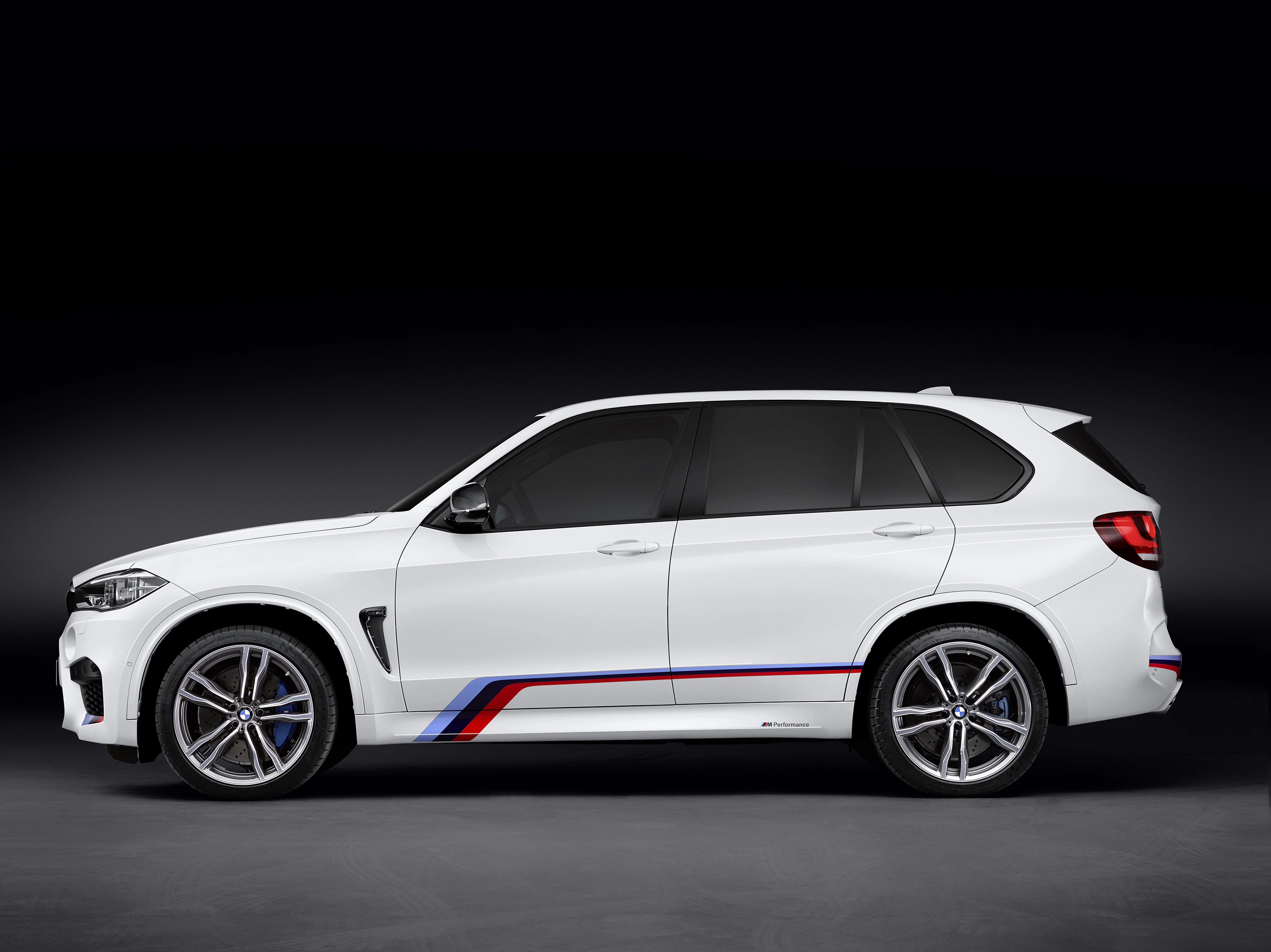 Weißer SUV mit M-Streifen an der Seite, vor dunklem Hintergrund. Sportliches Design mit blauen Bremssätteln und roten Akzenten.