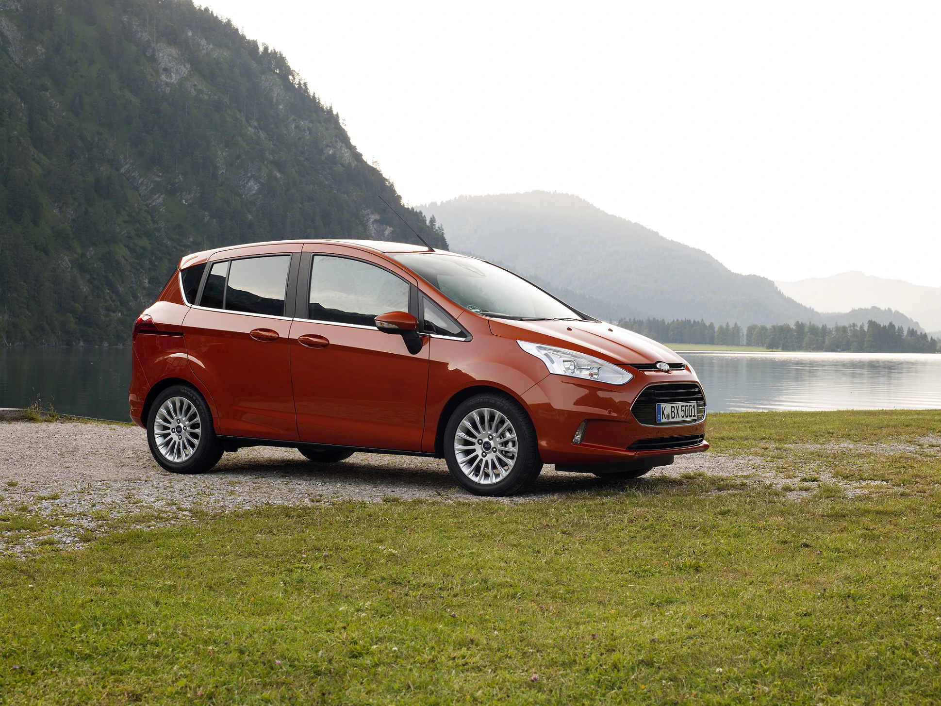 Roter Minivan parkt auf einer Wiese vor einem Bergsee, umgeben von grünen Hügeln und bewaldeten Bergen unter bewölktem Himmel.