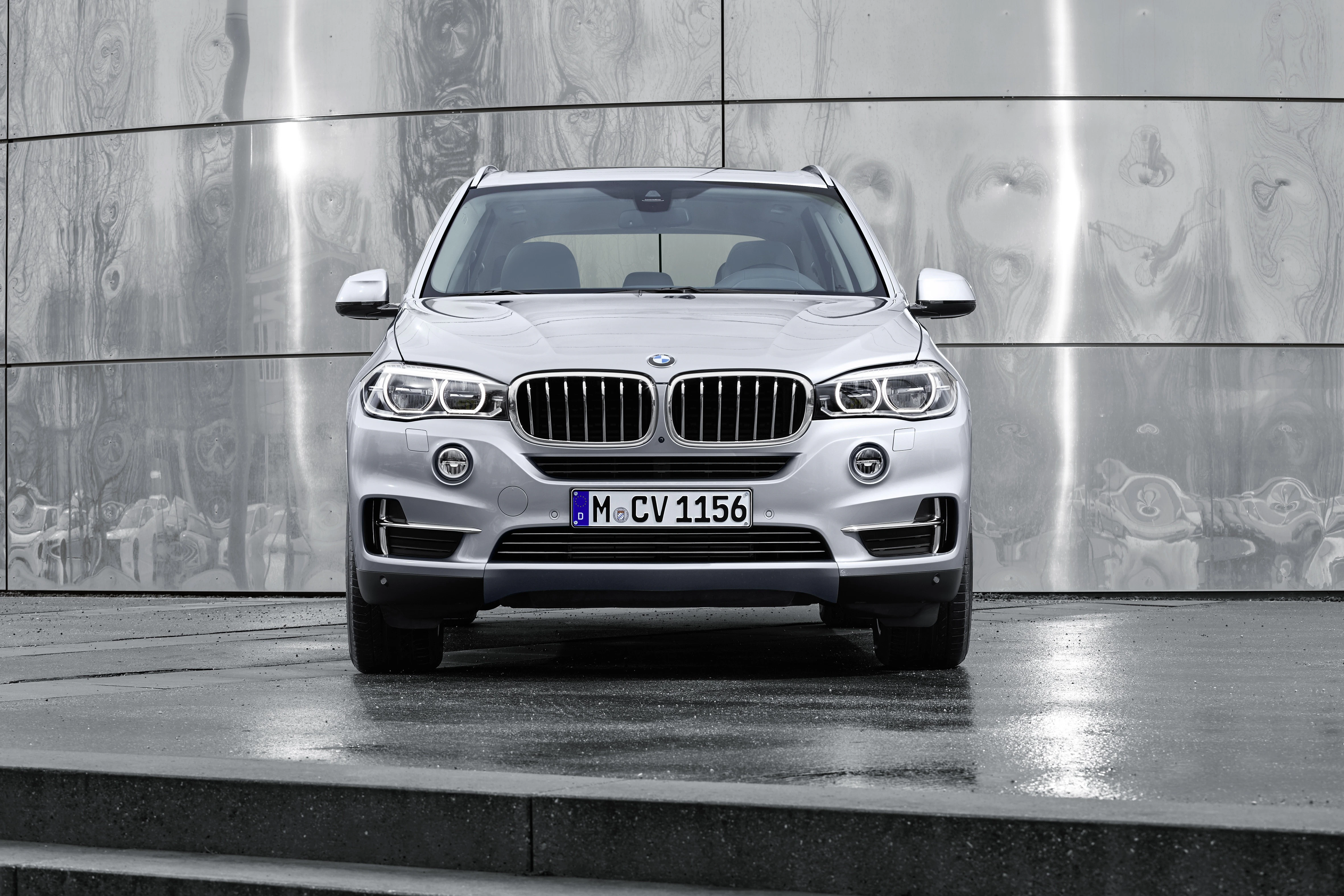 Silberner BMW-SUV, Frontansicht, auf nasser Straße vor einer modernen, metallischen Wand geparkt.