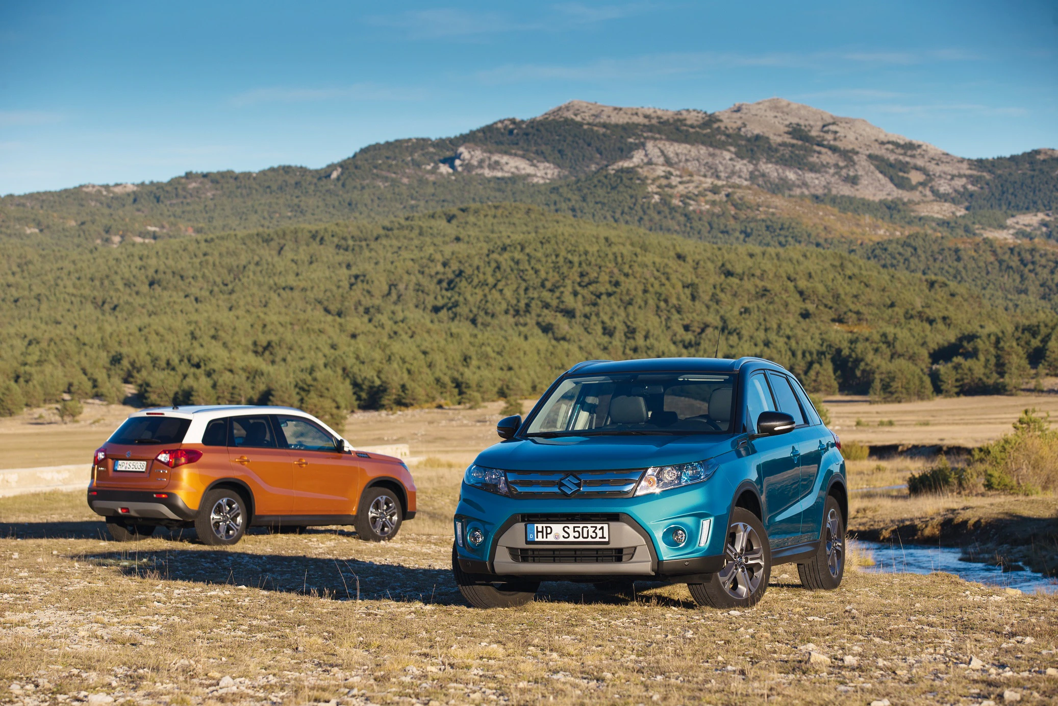 Zwei farbige SUVs parken in einer offenen Landschaft mit Bergen im Hintergrund unter blauem Himmel.