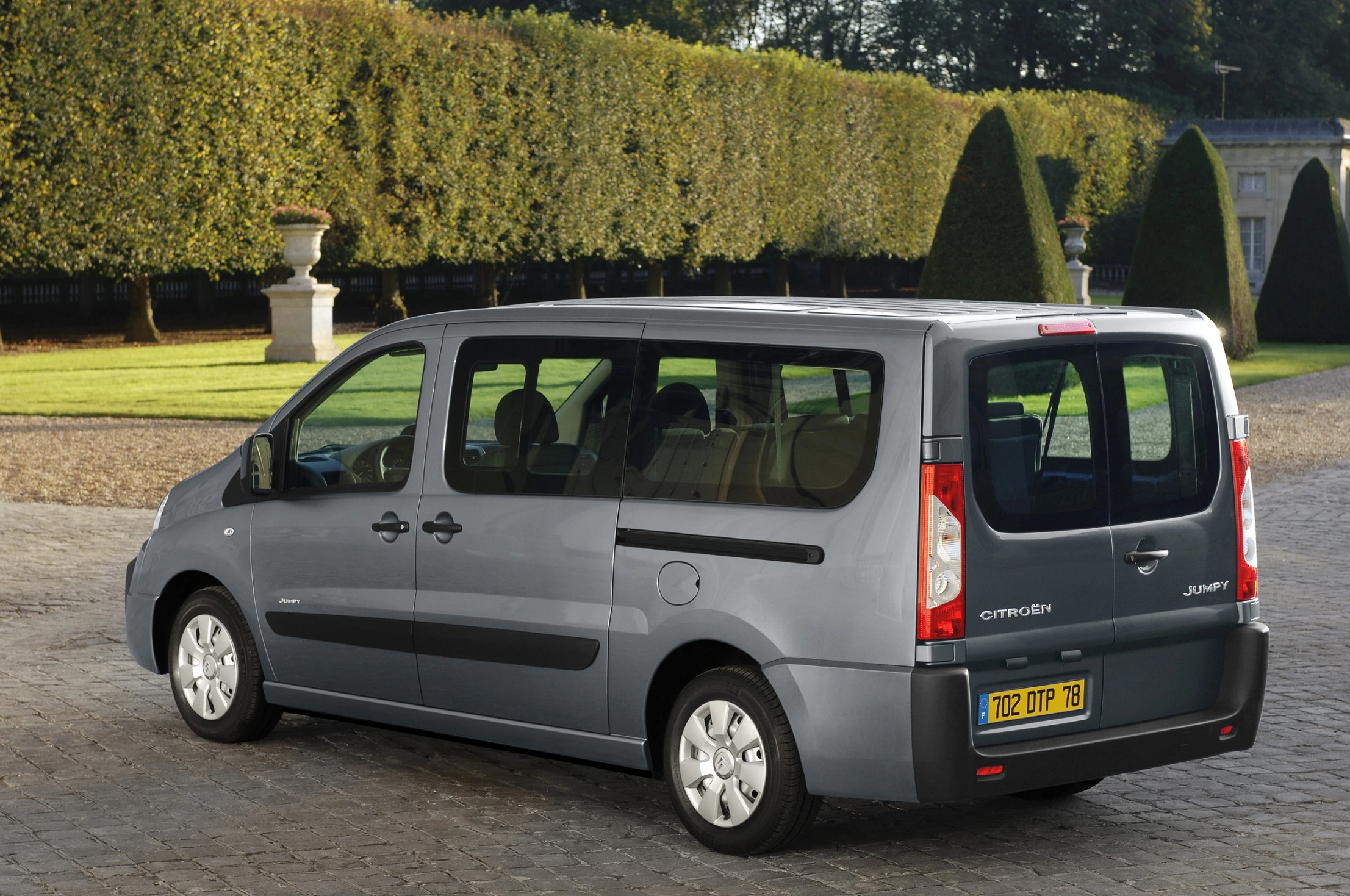 Silberner Citroën Jumpy Van, geparkt auf einem gepflasterten Weg, umgeben von gepflegten Hecken und einem großen Baum im Hintergrund.