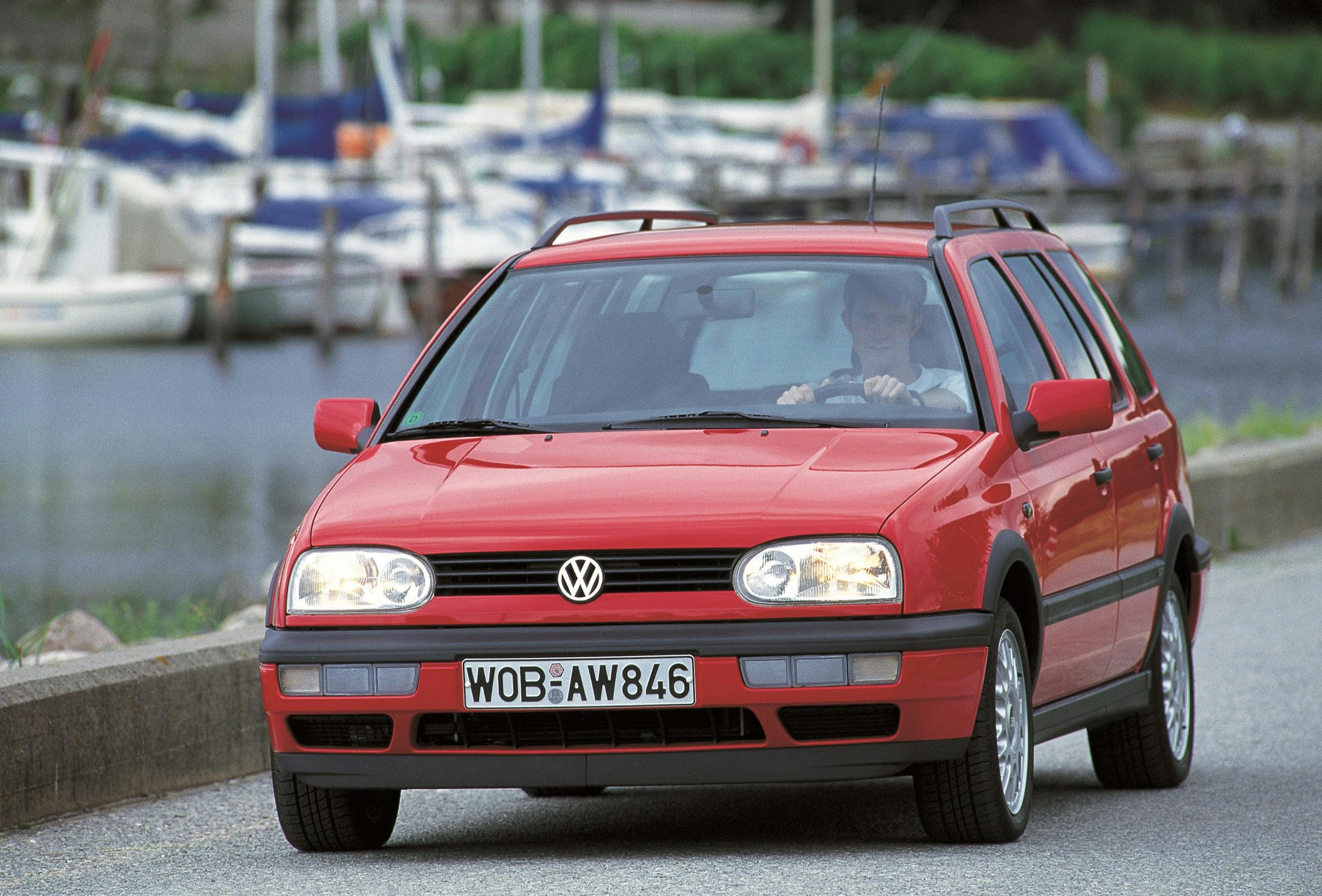 Roter Volkswagen Kombi fährt auf einer Straße am Wasser entlang, im Hintergrund sind Boote zu sehen.