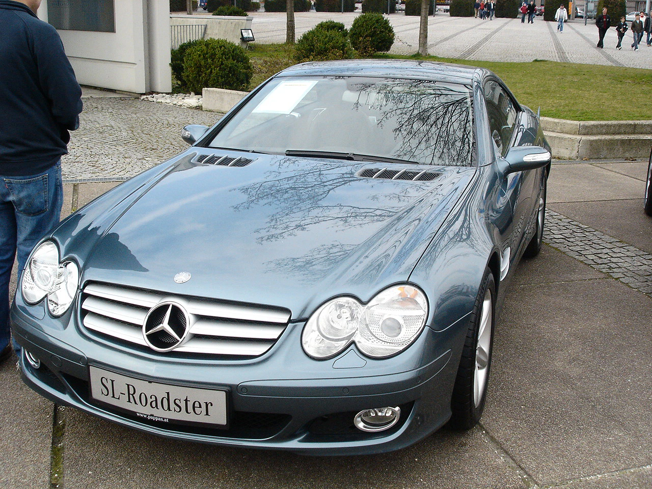 Silberner Mercedes SL-Roadster auf einem Parkplatz, umgeben von Gehwegen und Rasenflächen. Vorderansicht des Wagens bei Tageslicht.