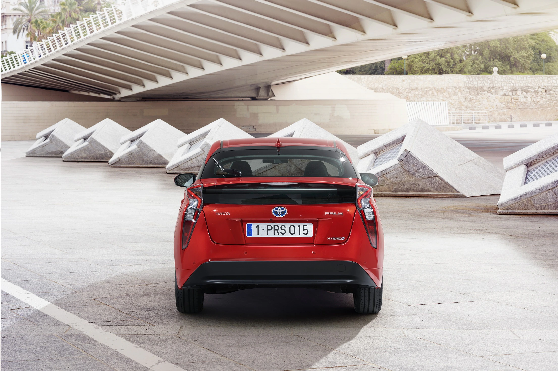 Rotes Auto steht auf gepflasterter Fläche unter modernem Brückenbogen, mit futuristischen Skulpturen im Hintergrund.
