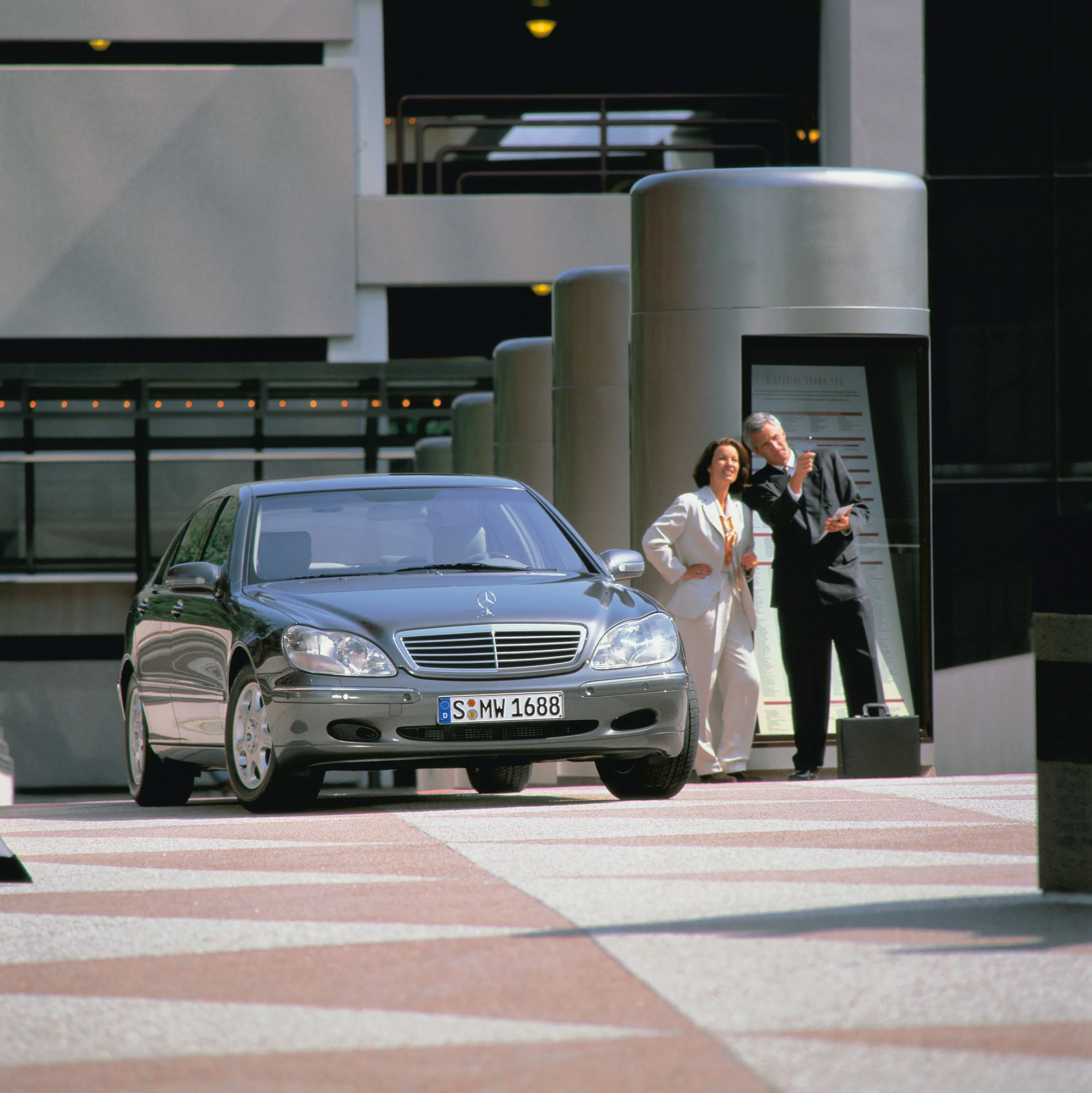 Silberner Mercedes-Benz in städtischem Ambiente, mit zwei elegant gekleideten Personen im Hintergrund, die auf etwas zu zeigen scheinen.
