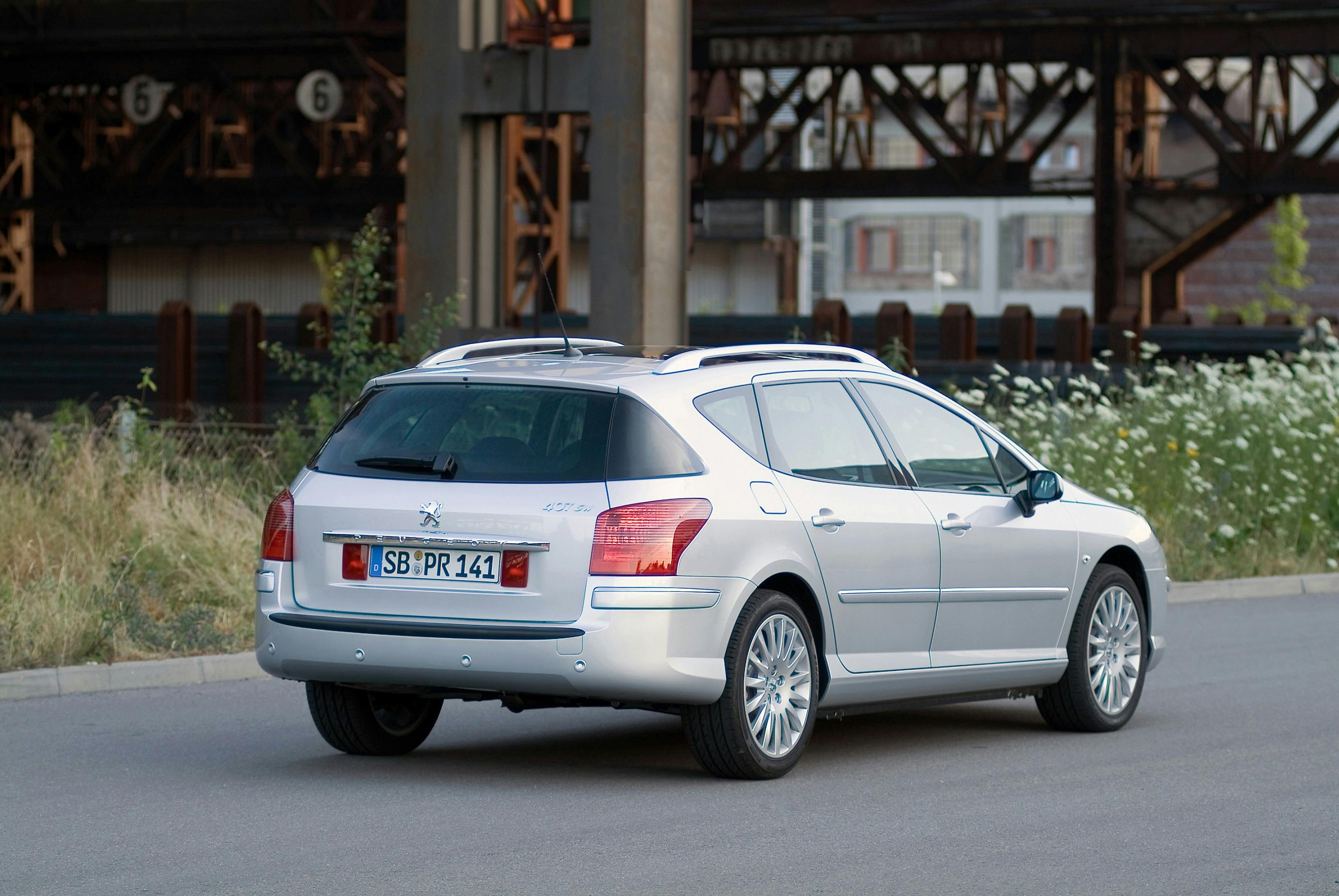 Silberner Kombi-Pkw steht vor einer Industrieanlage auf einer Straße, umgeben von Gras und Blumen.