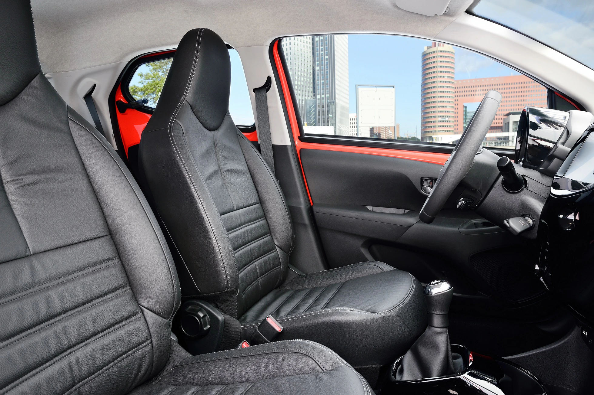 Innenansicht eines Autos mit schwarzen Ledersitzen, modernem Dashboard und Aussicht auf eine städtische Skyline durch das Fenster.