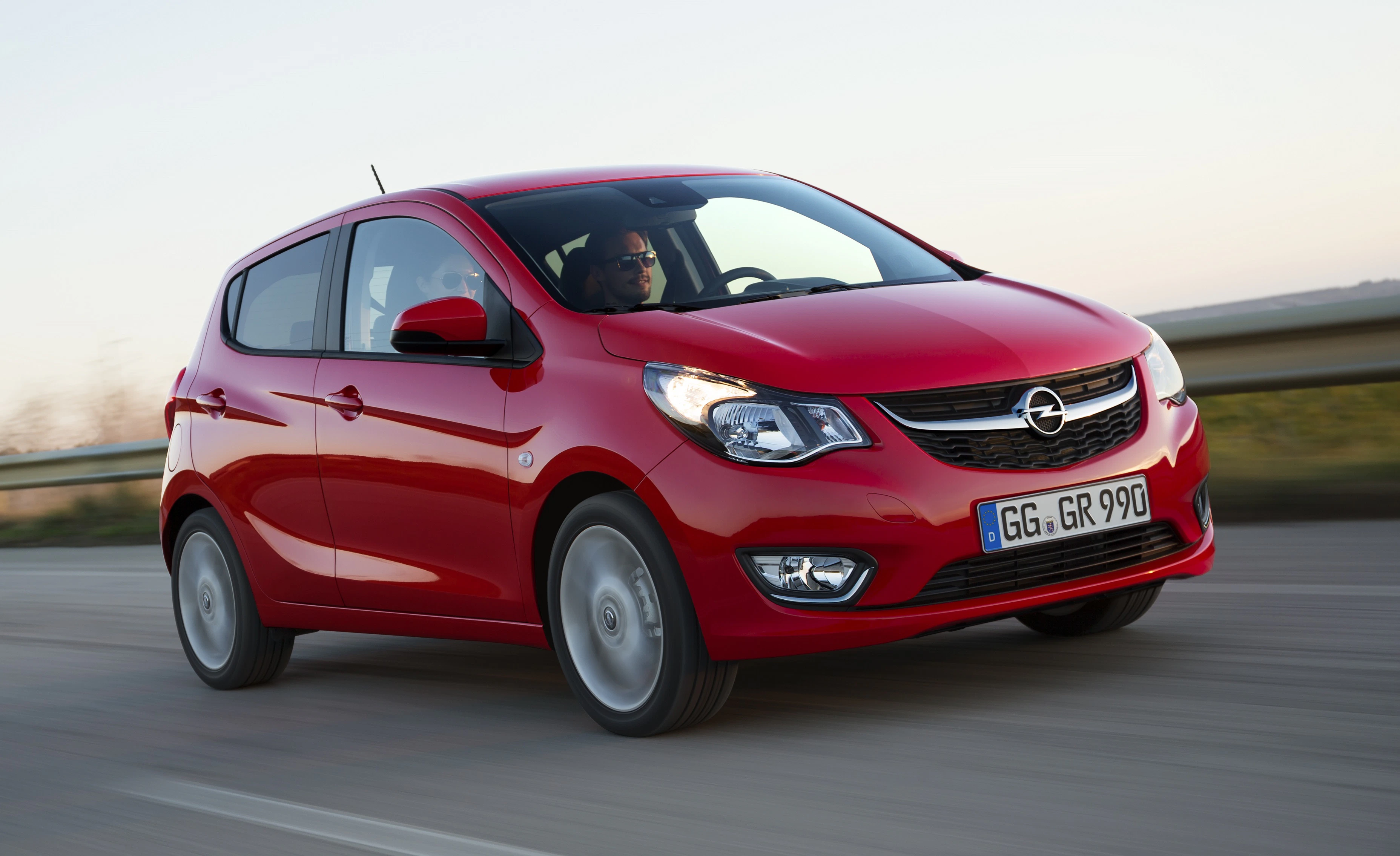 Roter Opel Karl fährt auf einer Straße, Fahrer am Steuer, Hintergrund verschwommen.
