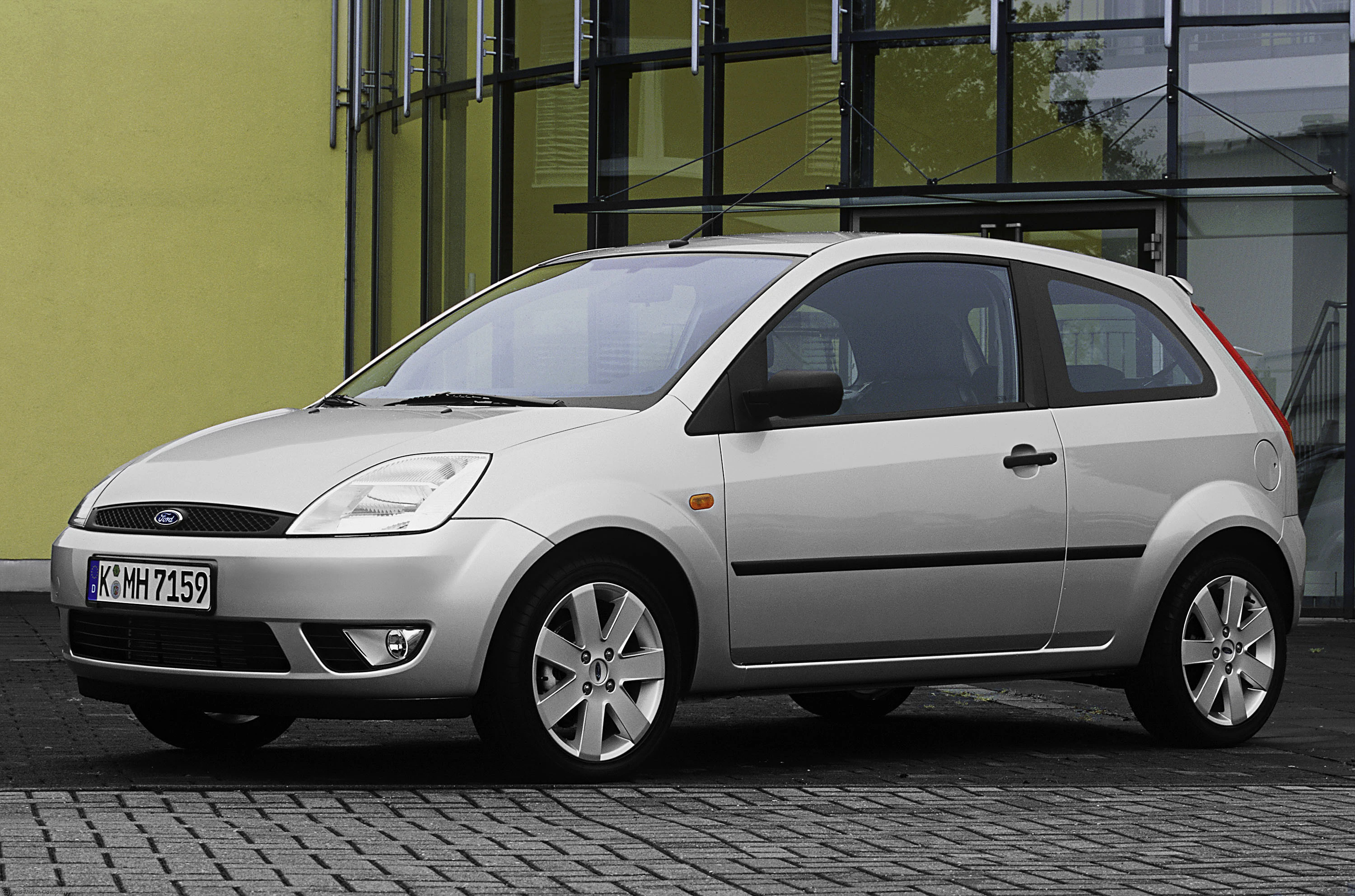 Silberner Ford Fiesta, geparkt vor einem modernen Gebäude mit Glasfassade auf gepflasterter Fläche.