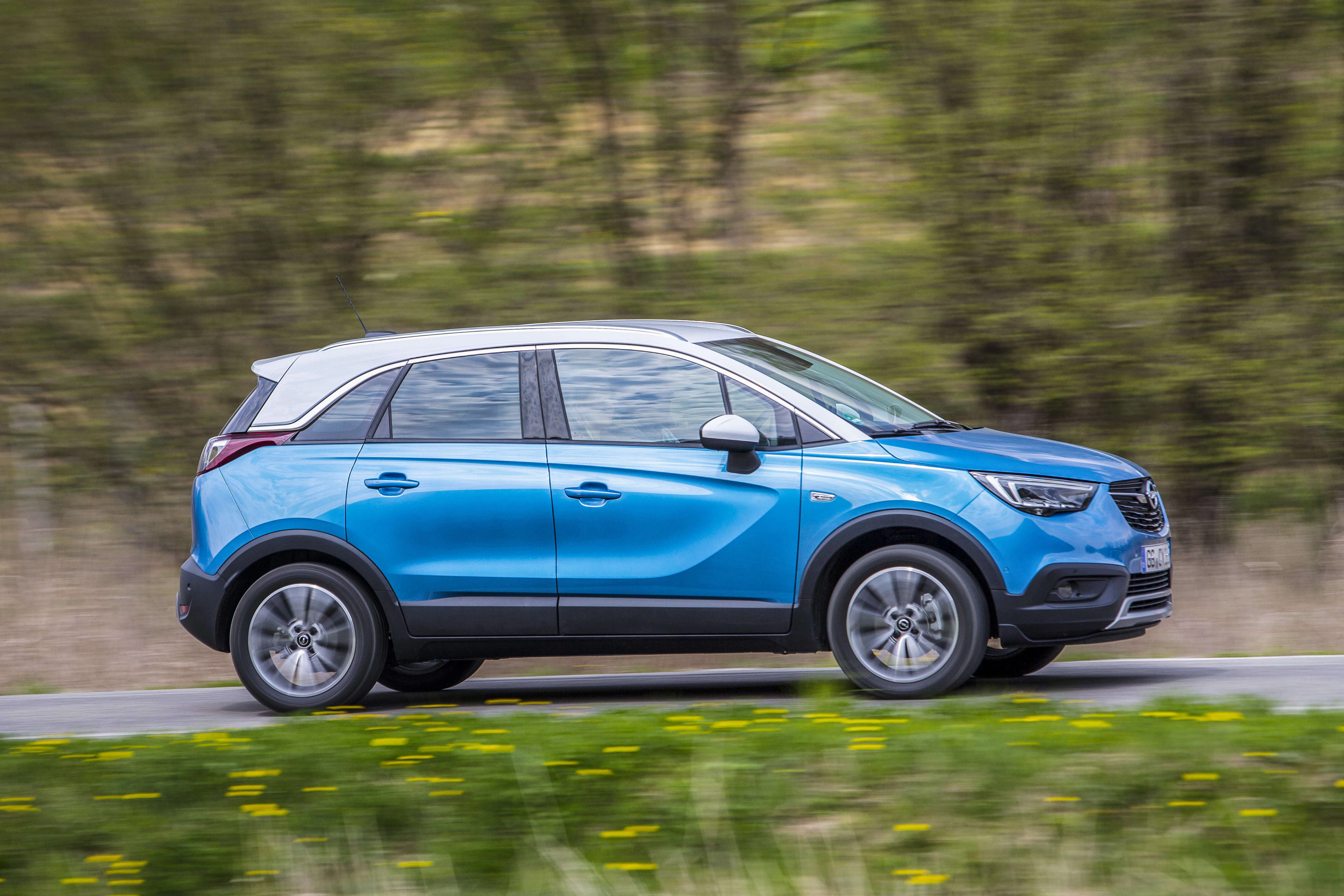 Blauer SUV fährt dynamisch auf einer Landstraße, im Hintergrund unscharfe grüne Vegetation. Kann als Opel Crossland bezeichnet werden.
