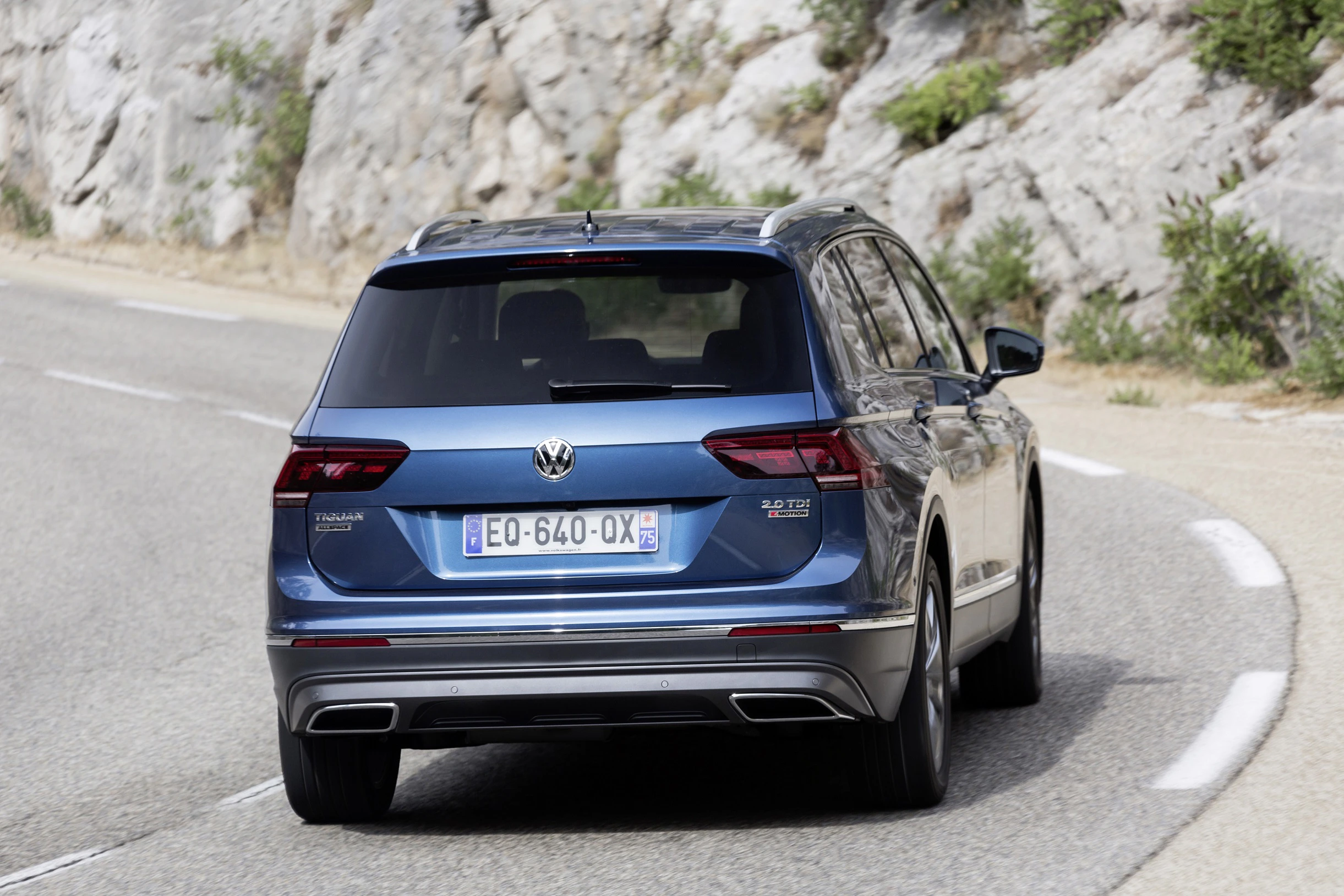 Ein blauer SUV fährt auf einer kurvigen Straße, umgeben von Felsen und Vegetation. Das Fahrzeug ist von hinten zu sehen.